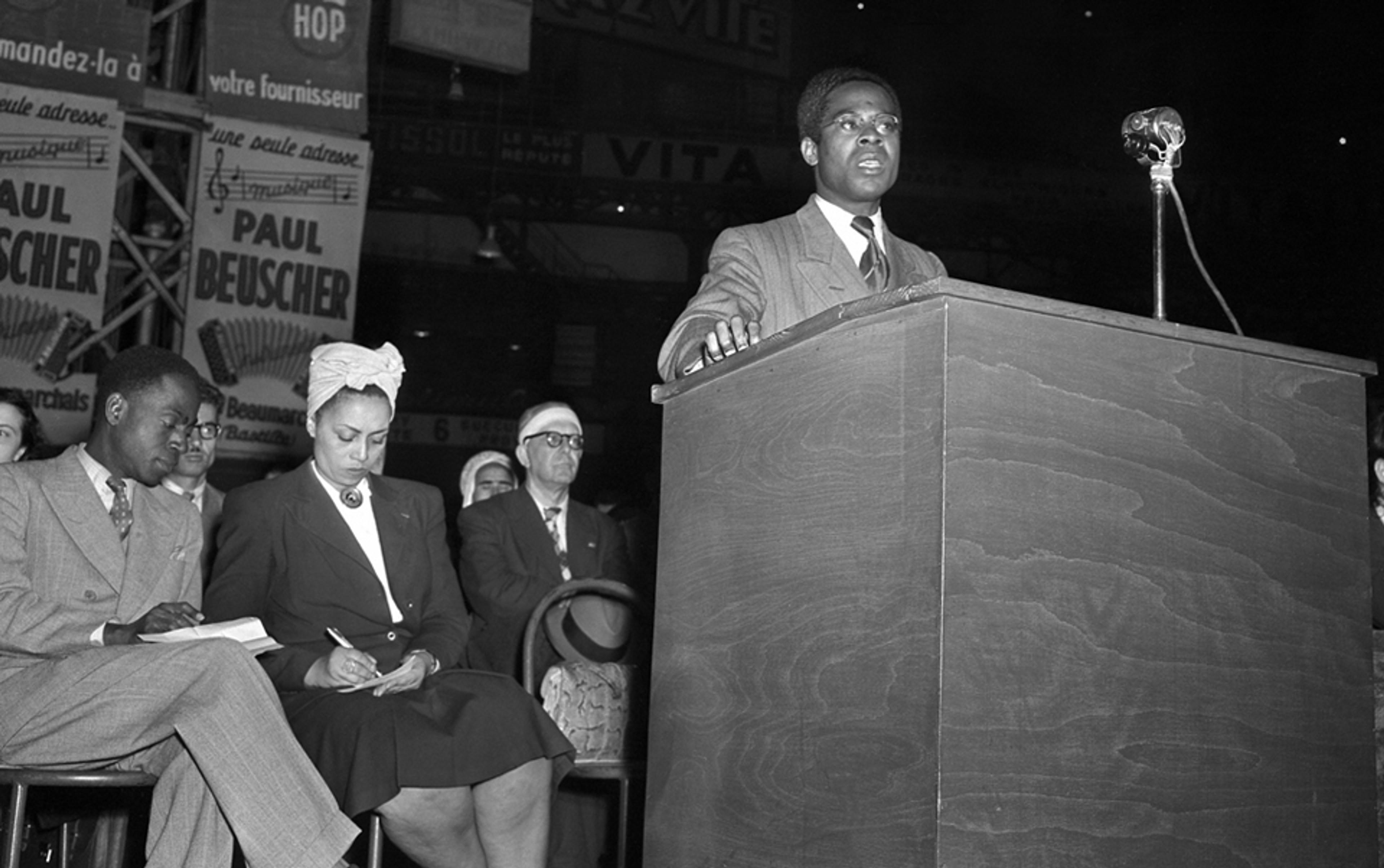 Black and white photo of a Black man in a suit speaking at a podium, with seated audience members including a Black woman writing in a notebook.