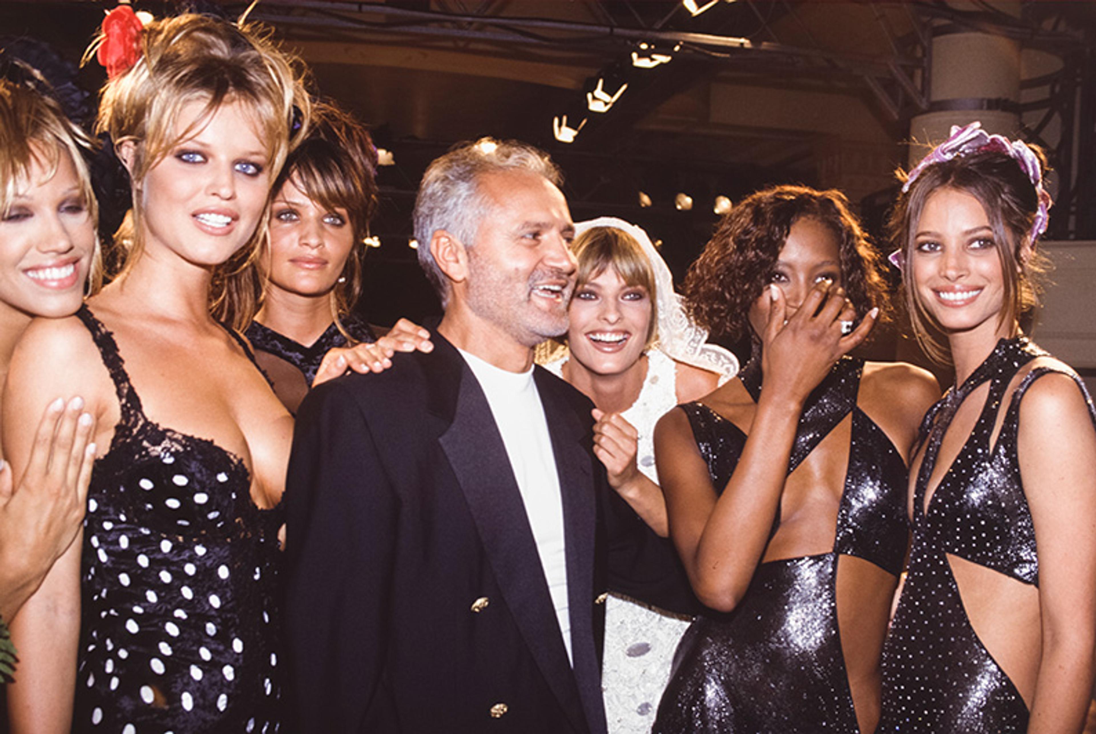 A smiling man in a suit surrounded by six women in glamorous outfits at an indoor event with bright overhead lighting.