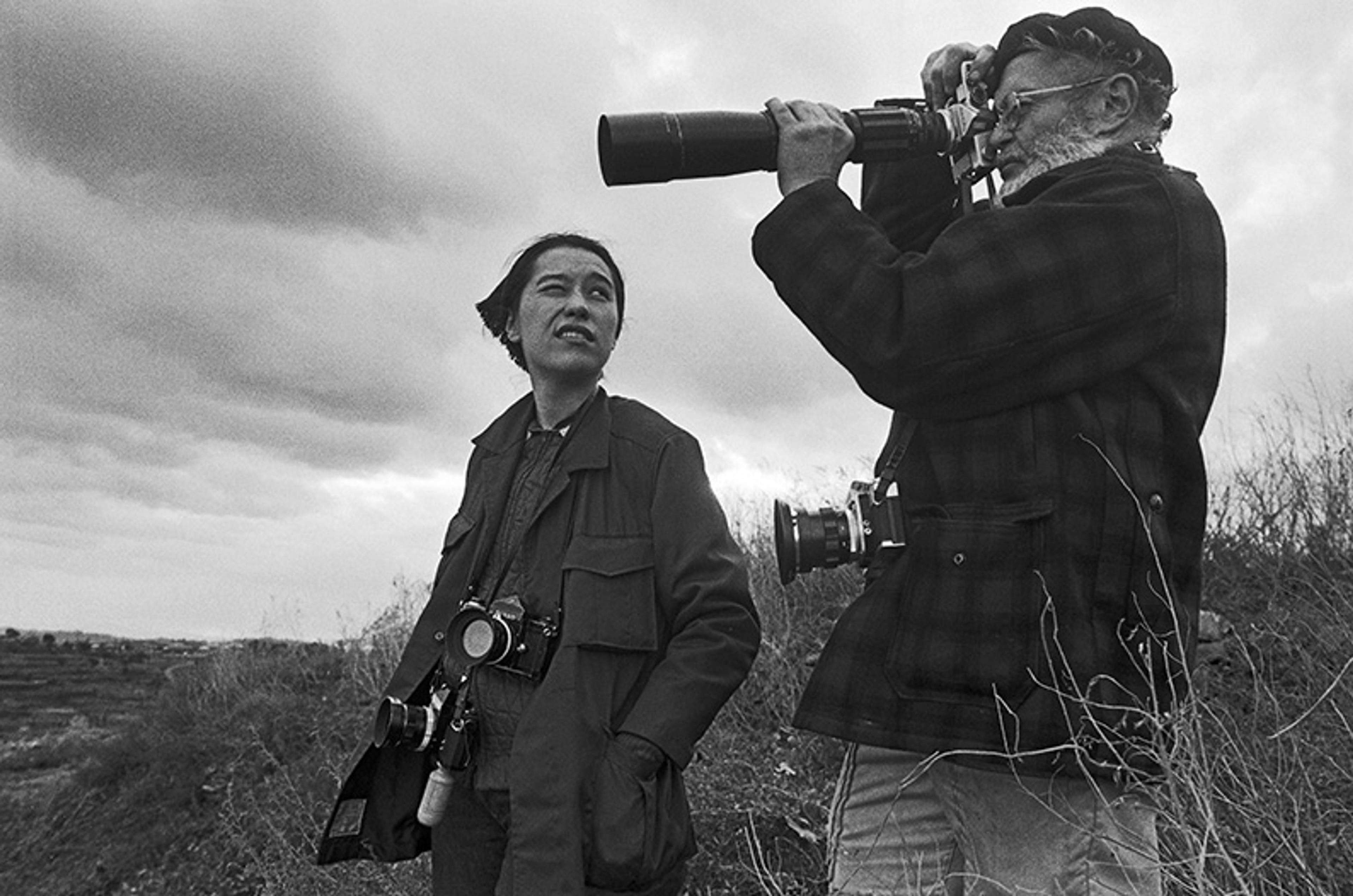 Black and white photo of two photographers outdoors, the man is holding a long-lens camera up to his eye, while the woman is looking at him.