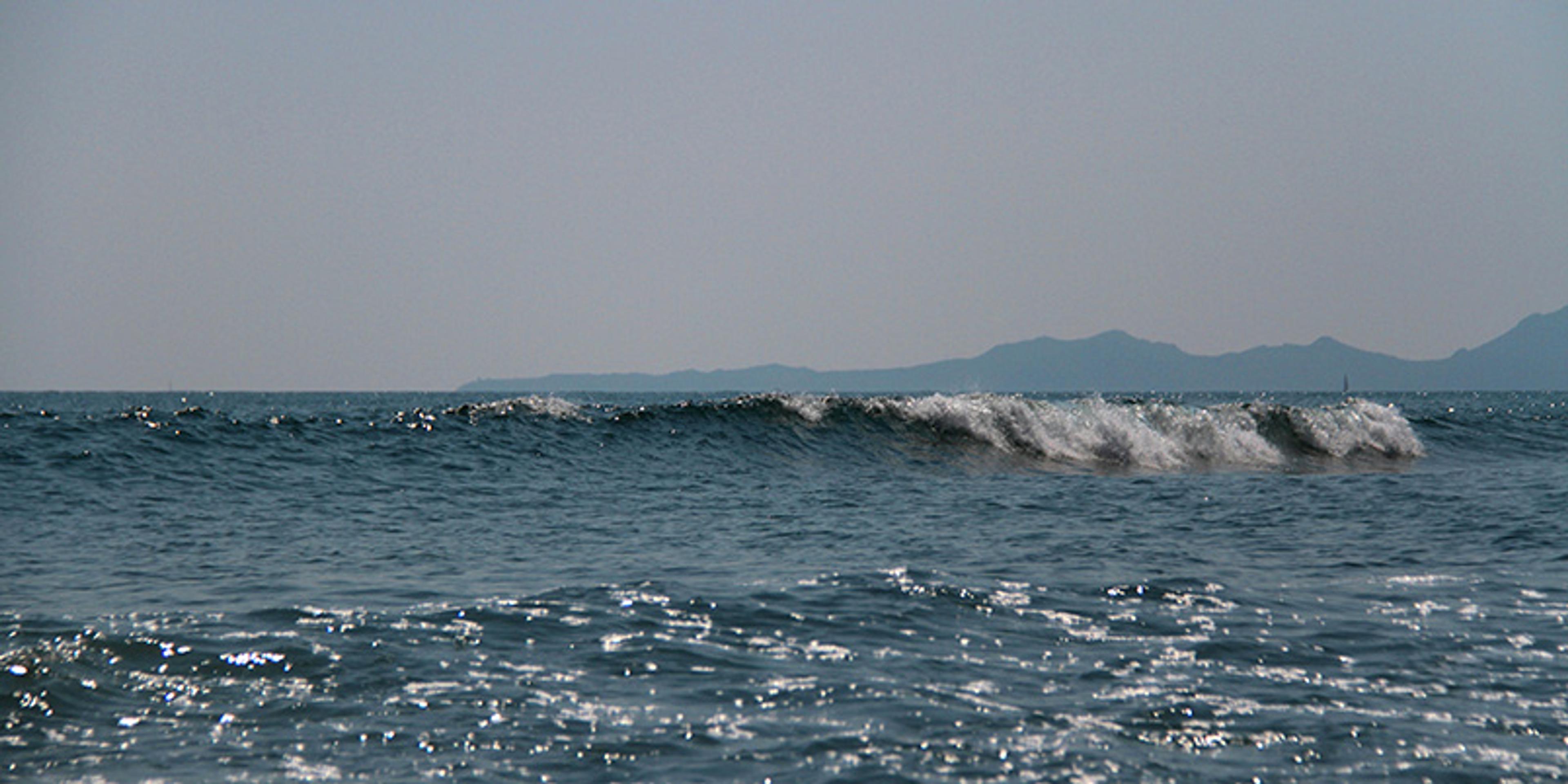 Photo of ocean waves under a clear sky with a distant mountain range visible on the horizon.