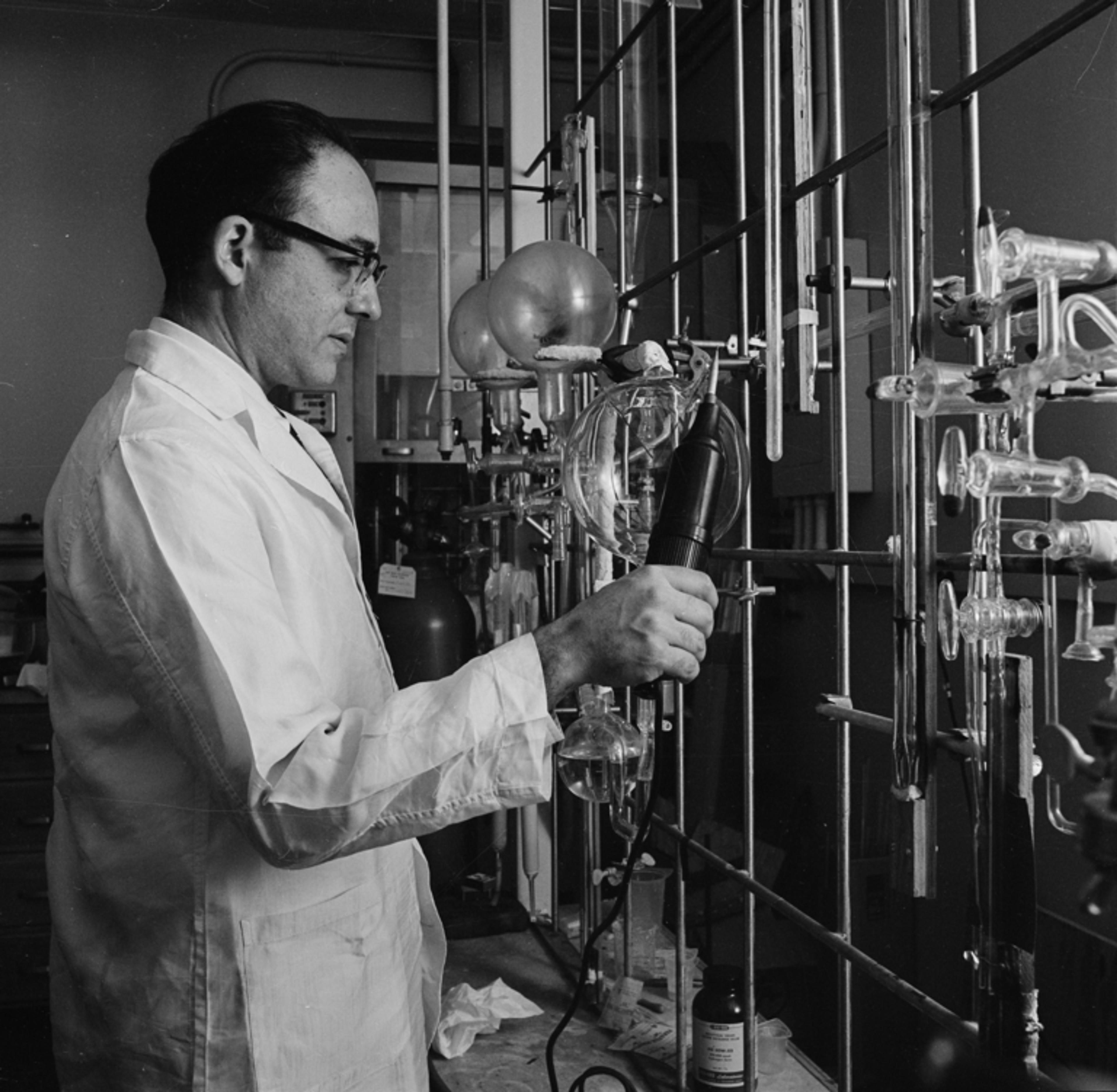 Black and white photo of a scientist in a lab coat using laboratory equipment amid glass beakers and tubes in a laboratory.