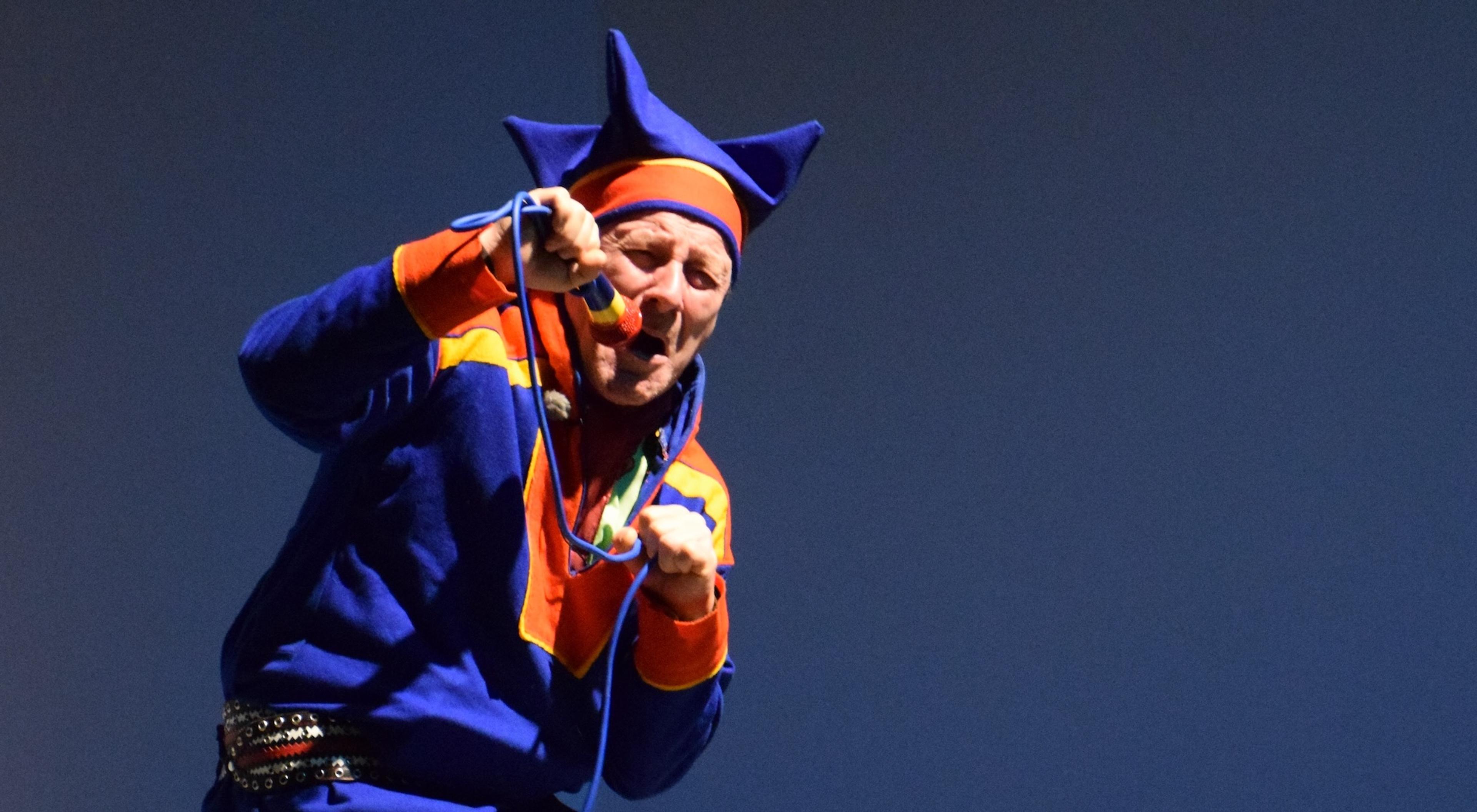 Photo of a performer in traditional Sami attire singing into a microphone on stage with a plain blue background.