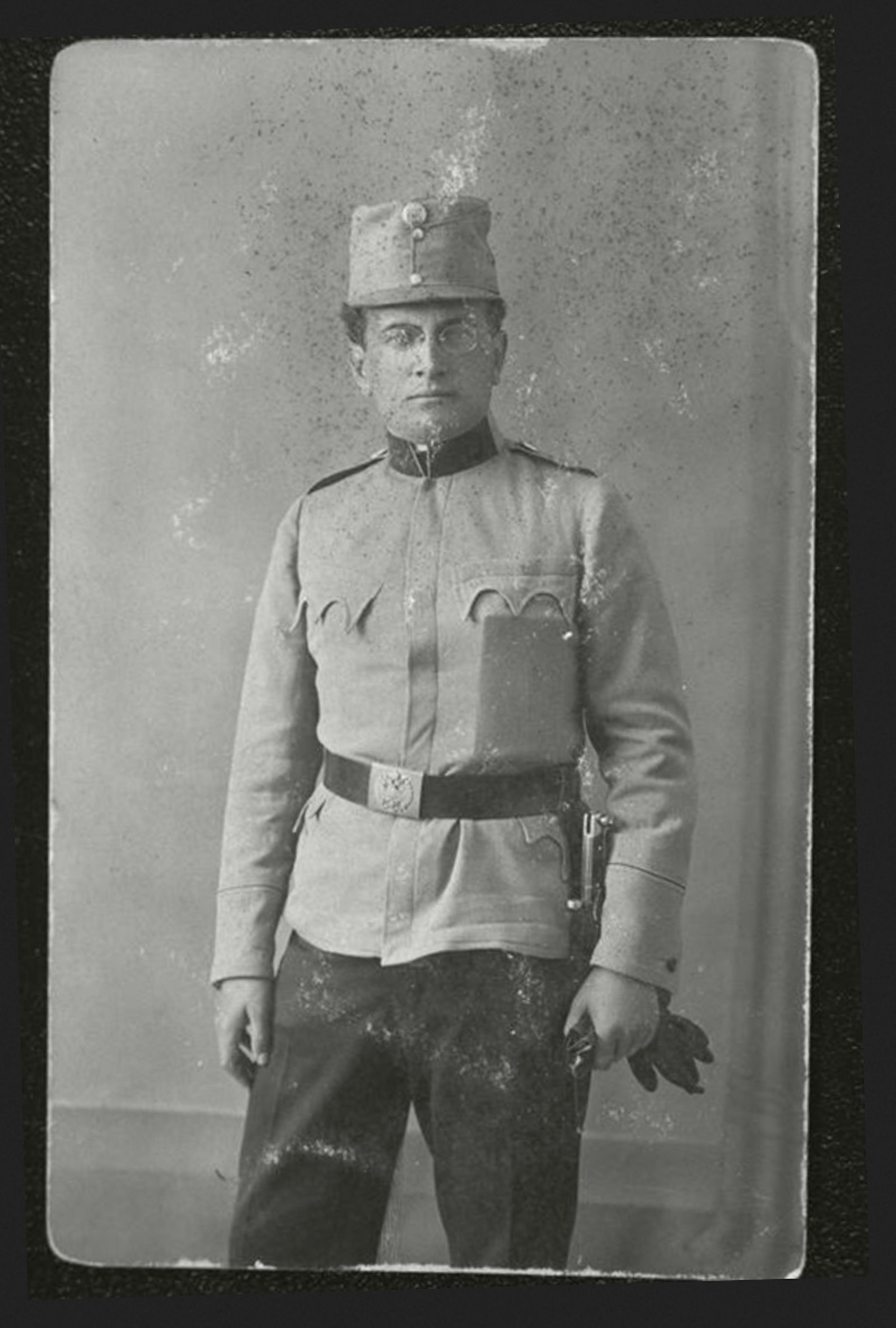 Black-and-white photo of a man in a military uniform with a hat and belt standing against a plain studio backdrop holding gloves.