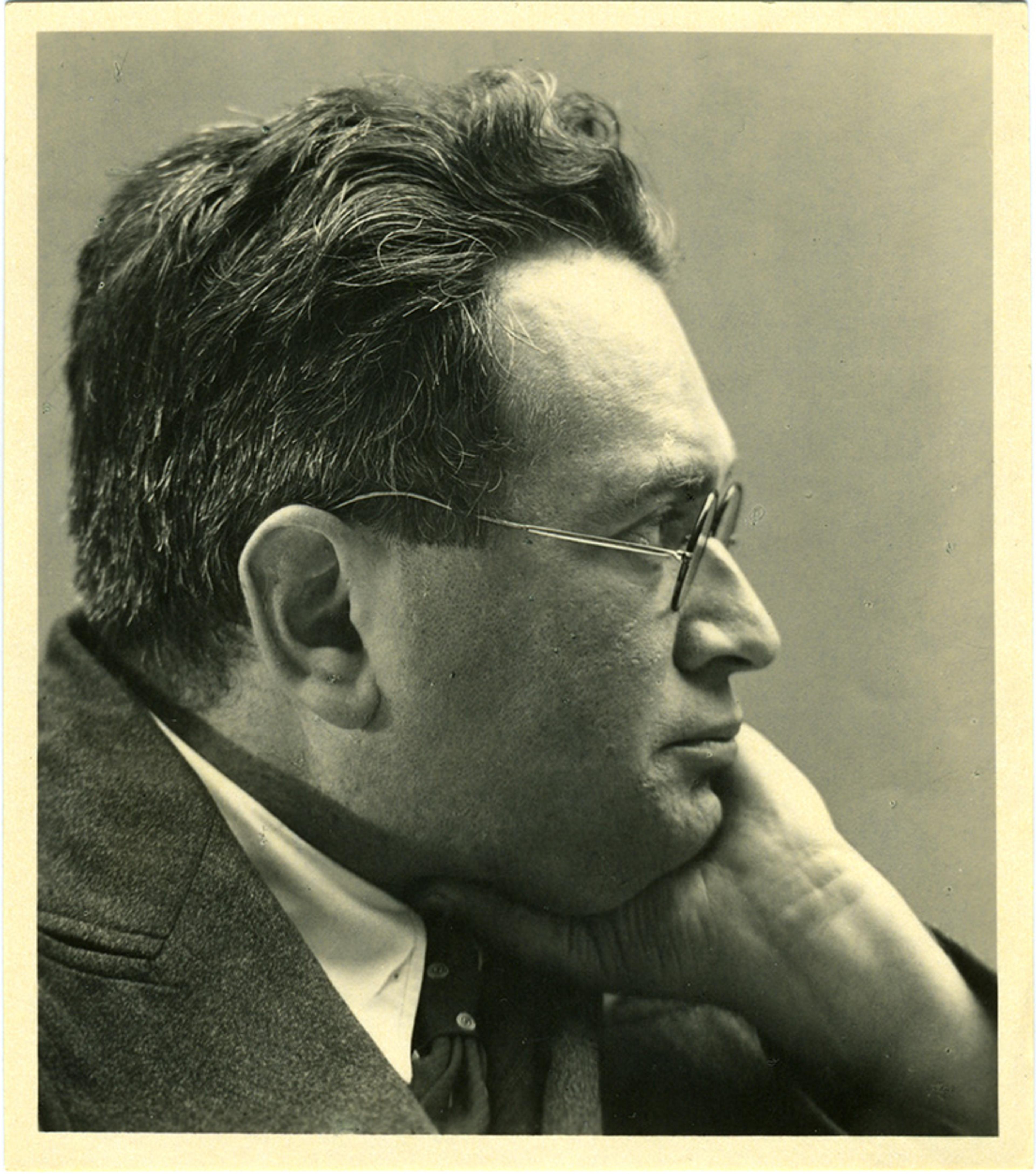 Black-and-white photo of a man in glasses with his chin resting on his hand, gazing to the right, dressed in a suit and tie.