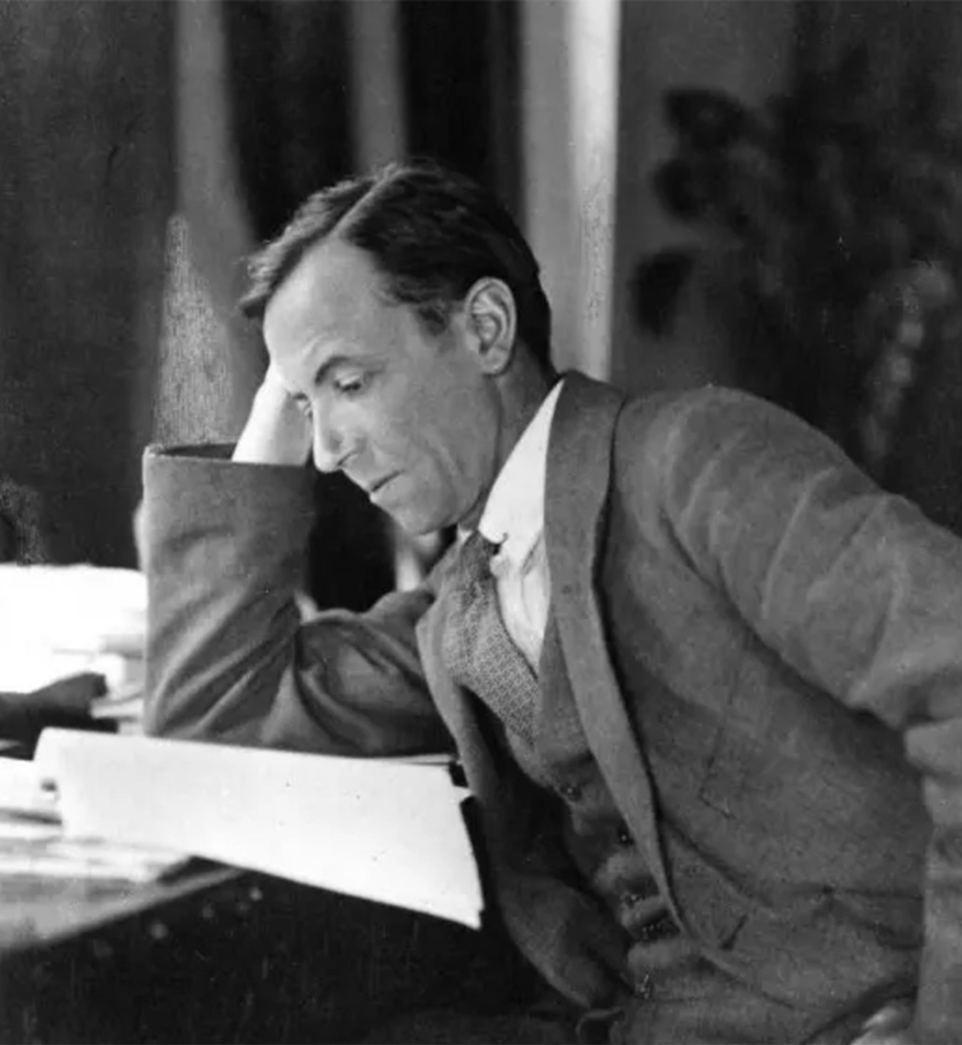 Black and white photo of a man in a suit sitting at a desk looking at papers with his head resting on his hand.