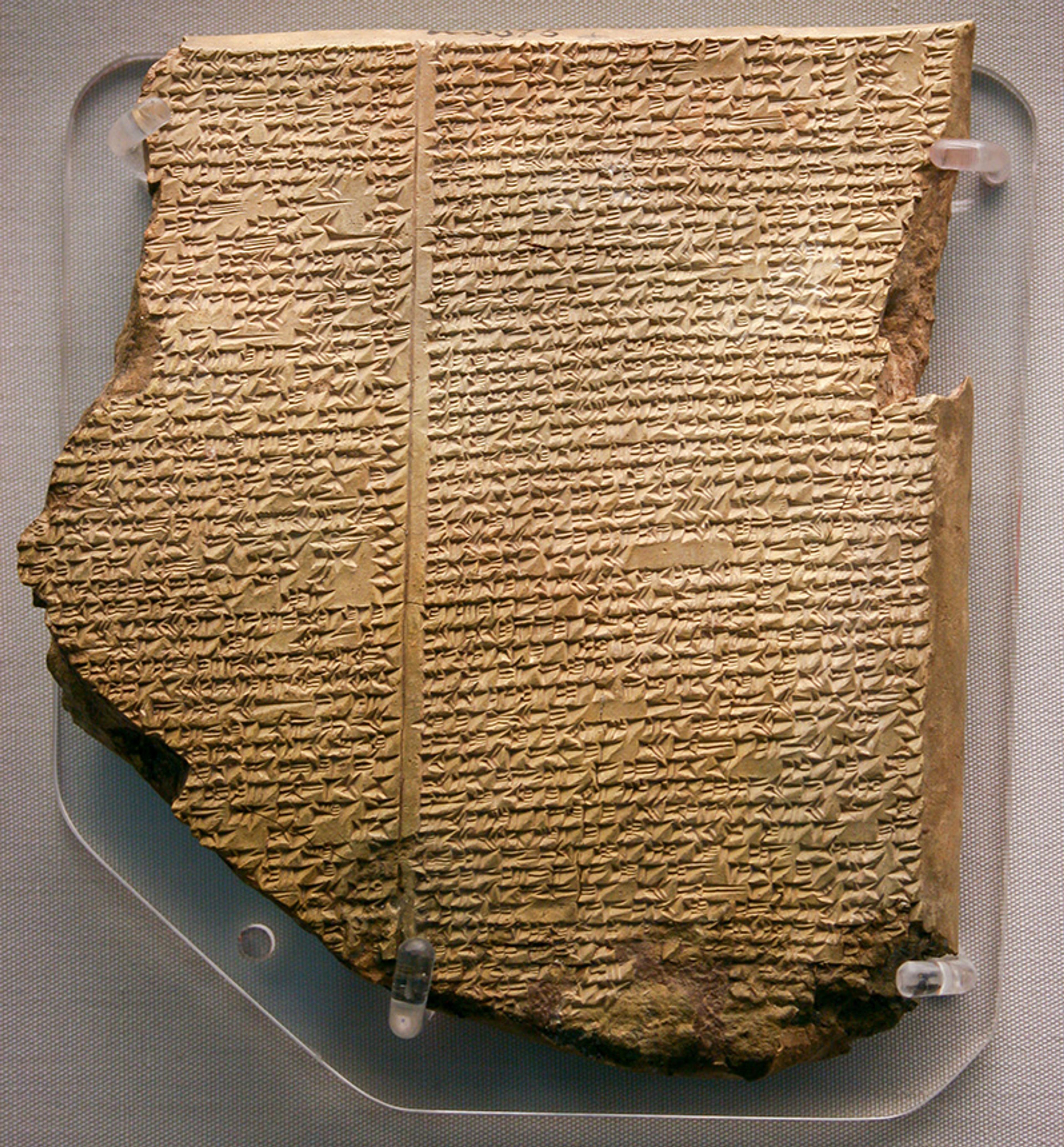 Photo of an ancient clay tablet with cuneiform script, displayed in a museum. The tablet is partially broken and mounted on a stand.