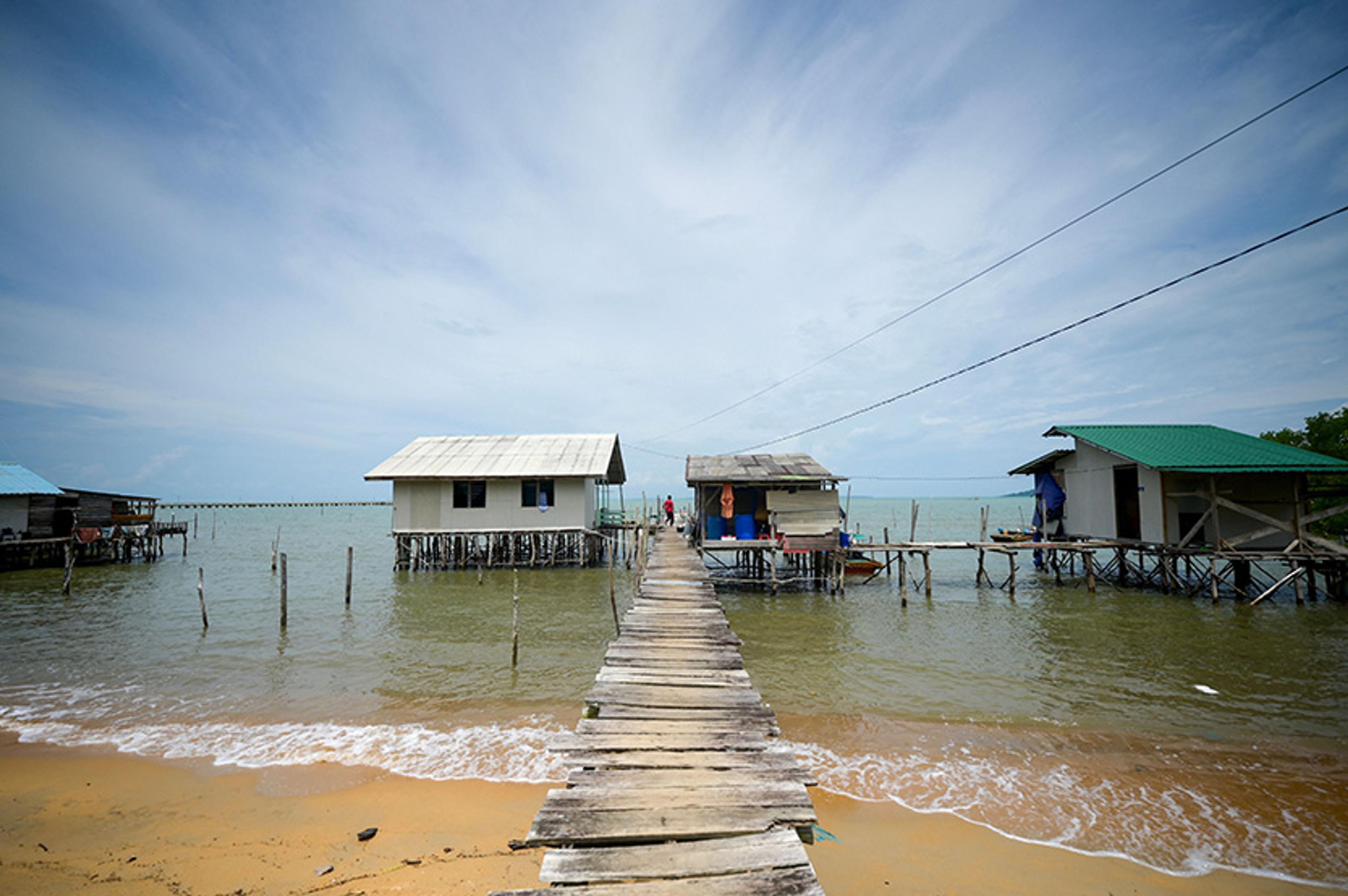 Photo of stilt houses over water connected by a wooden walkway, with a sandy beach and calm sea under a partly cloudy sky.