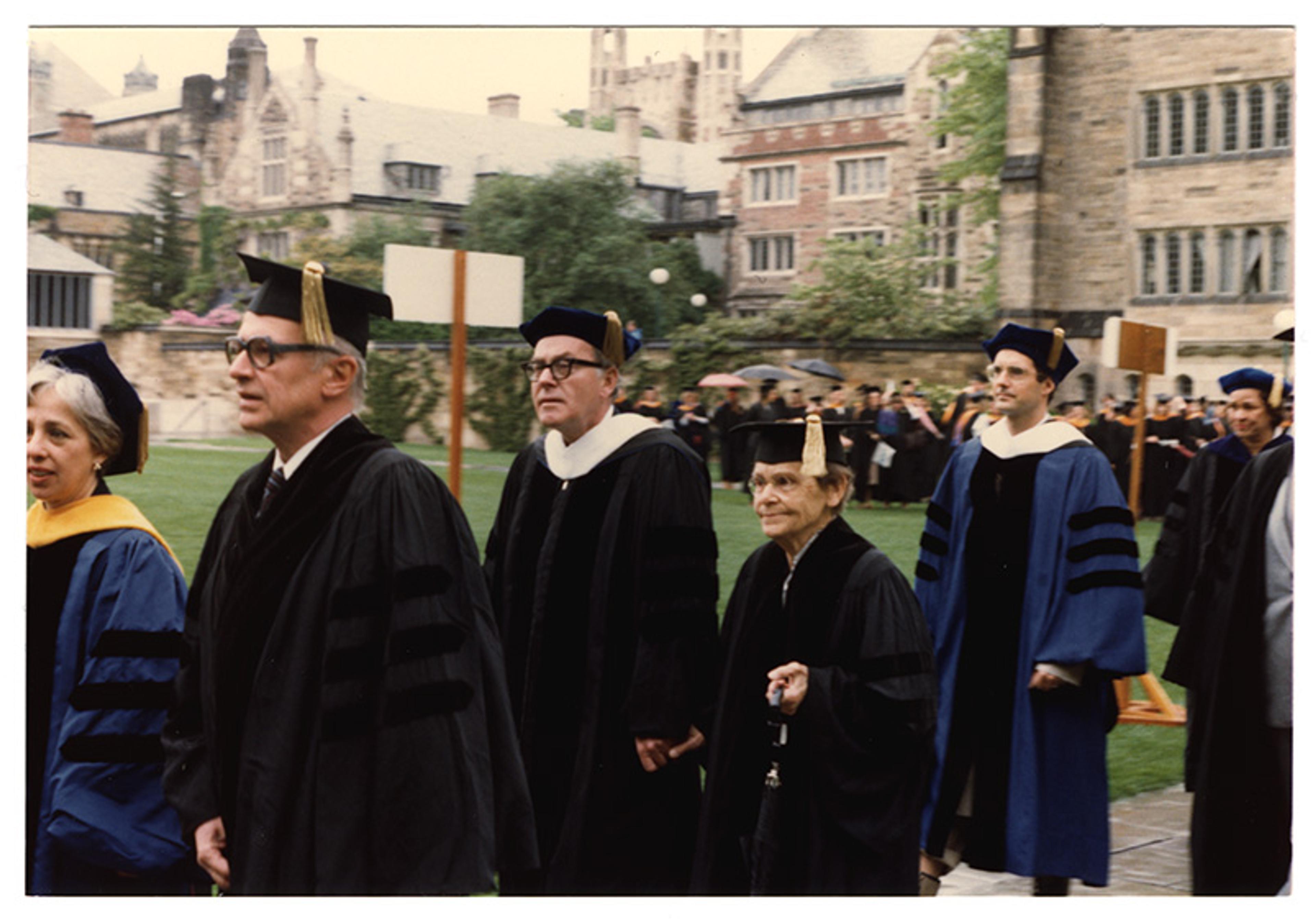 Photo of people in academic regalia at an outdoor graduation ceremony, with historic buildings in the background. Among them is an elderly Barbara McClintock.