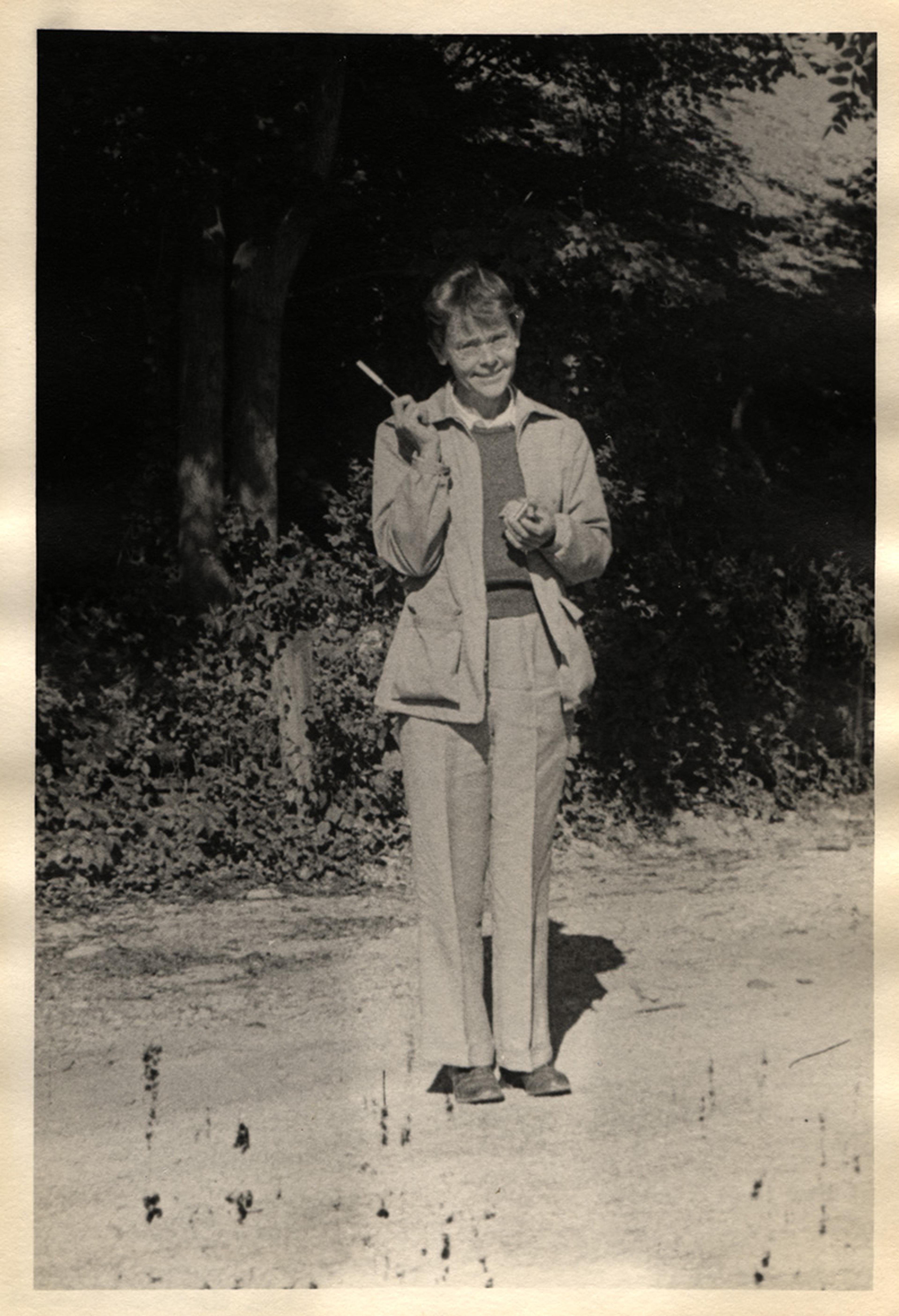 Black-and-white photo of a woman in trousers and jacket, smiling and holding a cigarette, with foliage in the background.