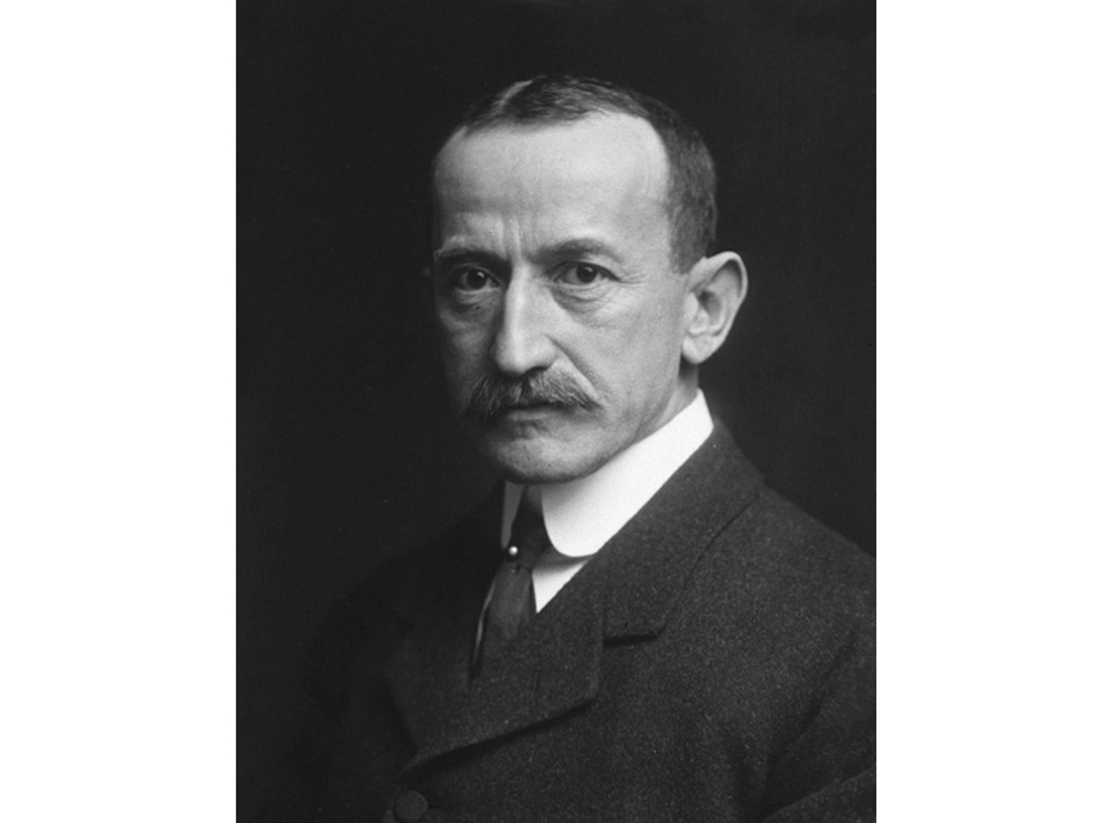 Black-and-white photo of a serious man with a moustache, wearing a formal suit and tie, against a dark background.