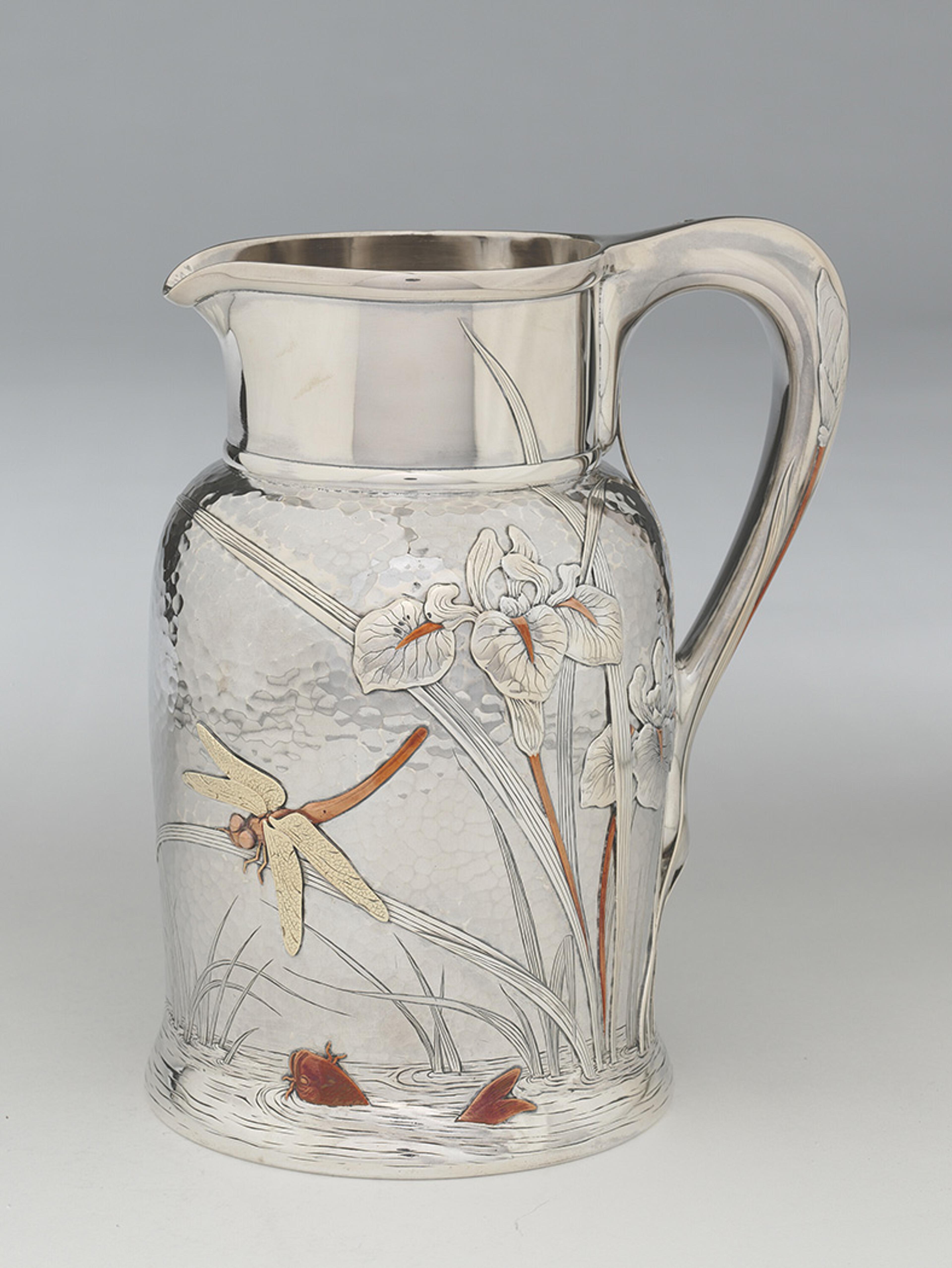 An ornate silver jug, featuring engraved and applied metal details of iris flowers, a dragonfly and a splashing carp fish.