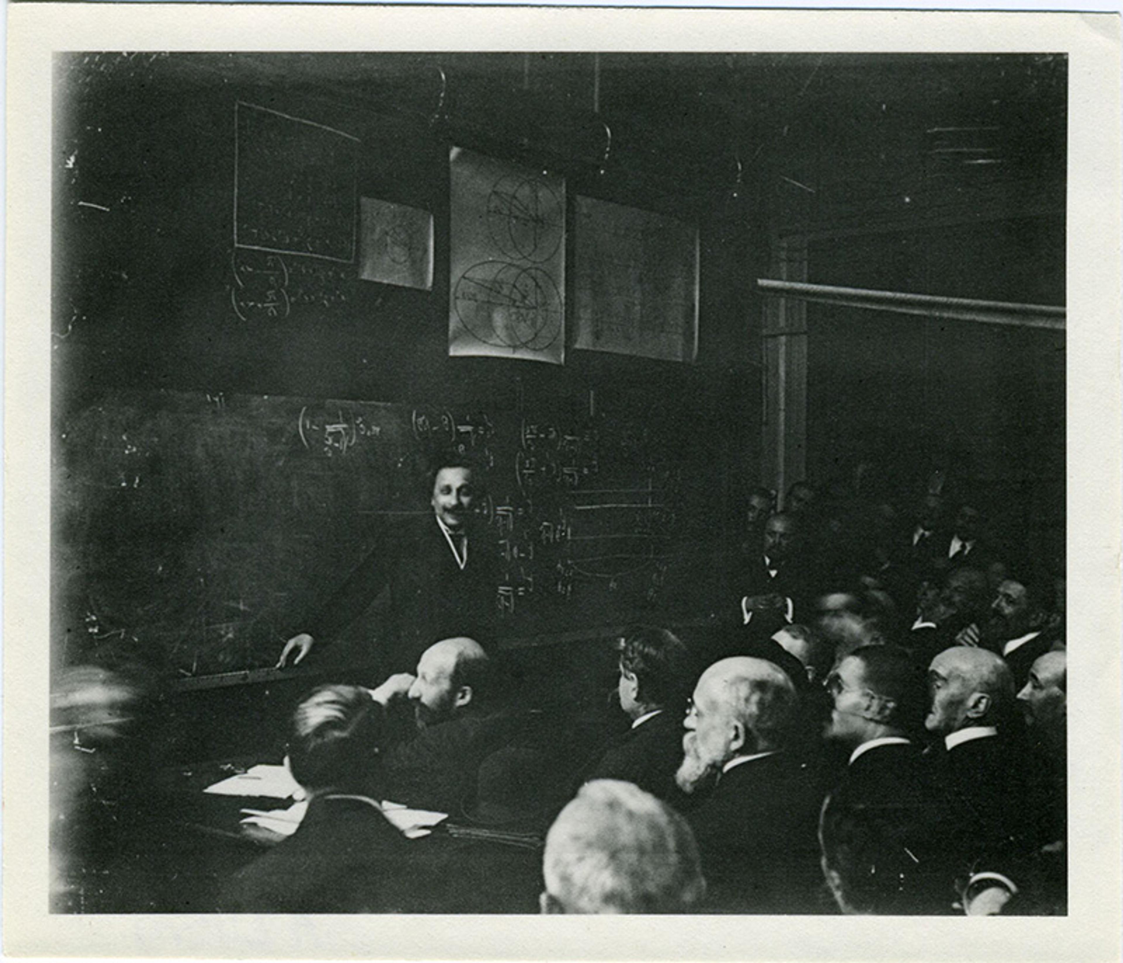 Black-and-white photo of a young Albert Einstein explaining equations on a chalkboard to an audience of seated men in a classroom.