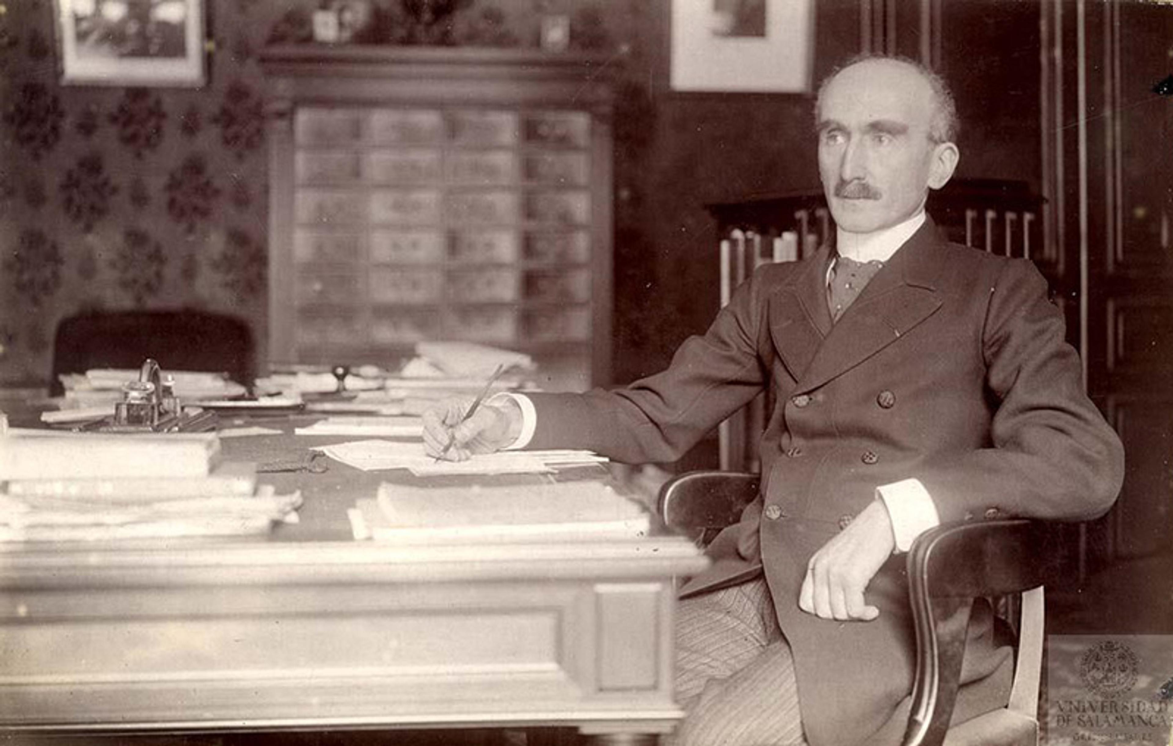 Black and white photo of a man in a suit sitting at a desk with papers, in an office with detailed wallpaper and framed pictures.