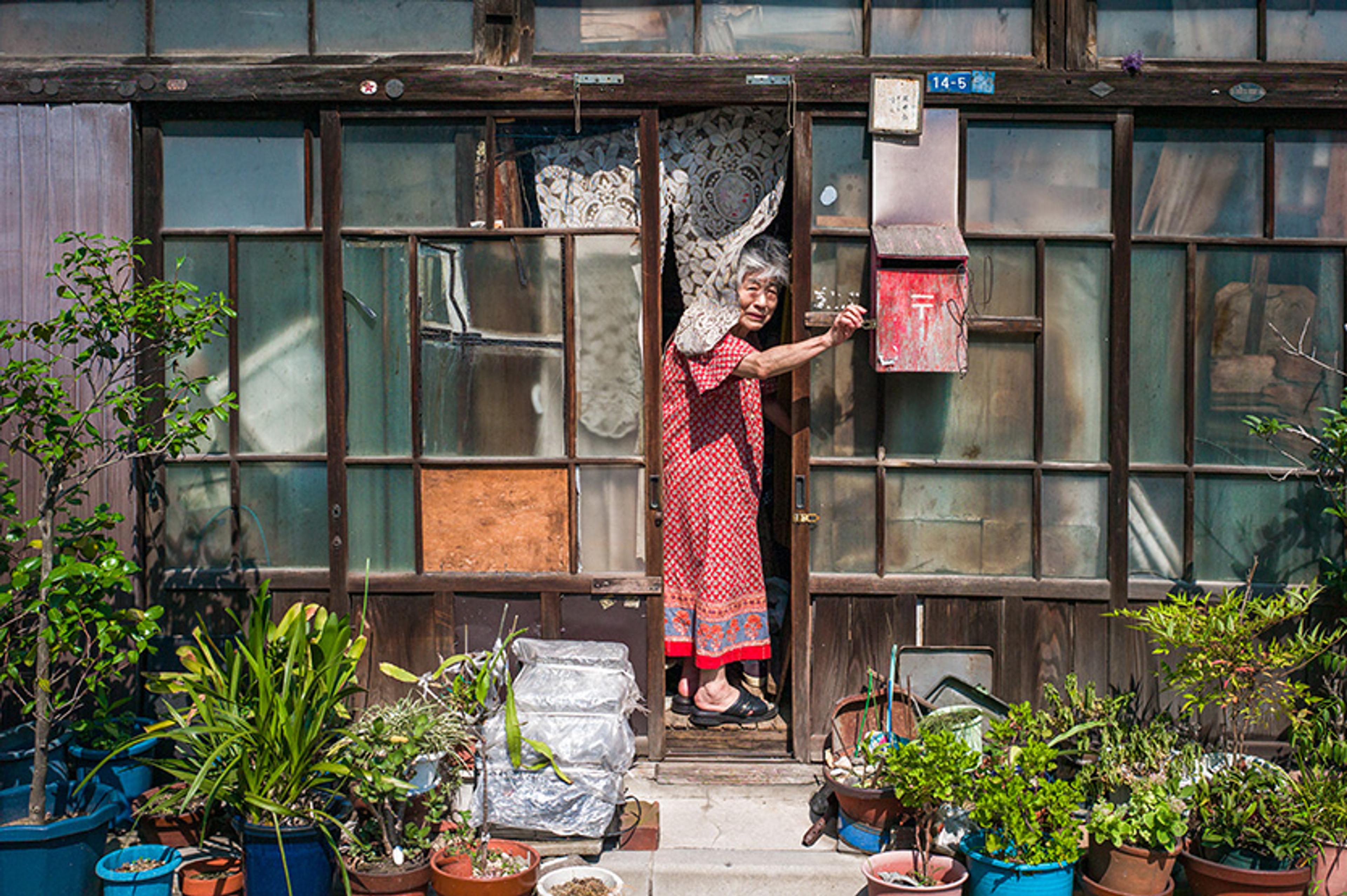 An elderly Japanese woman in a red dress standing at the entrance of a rustic house with numerous plants in pots.