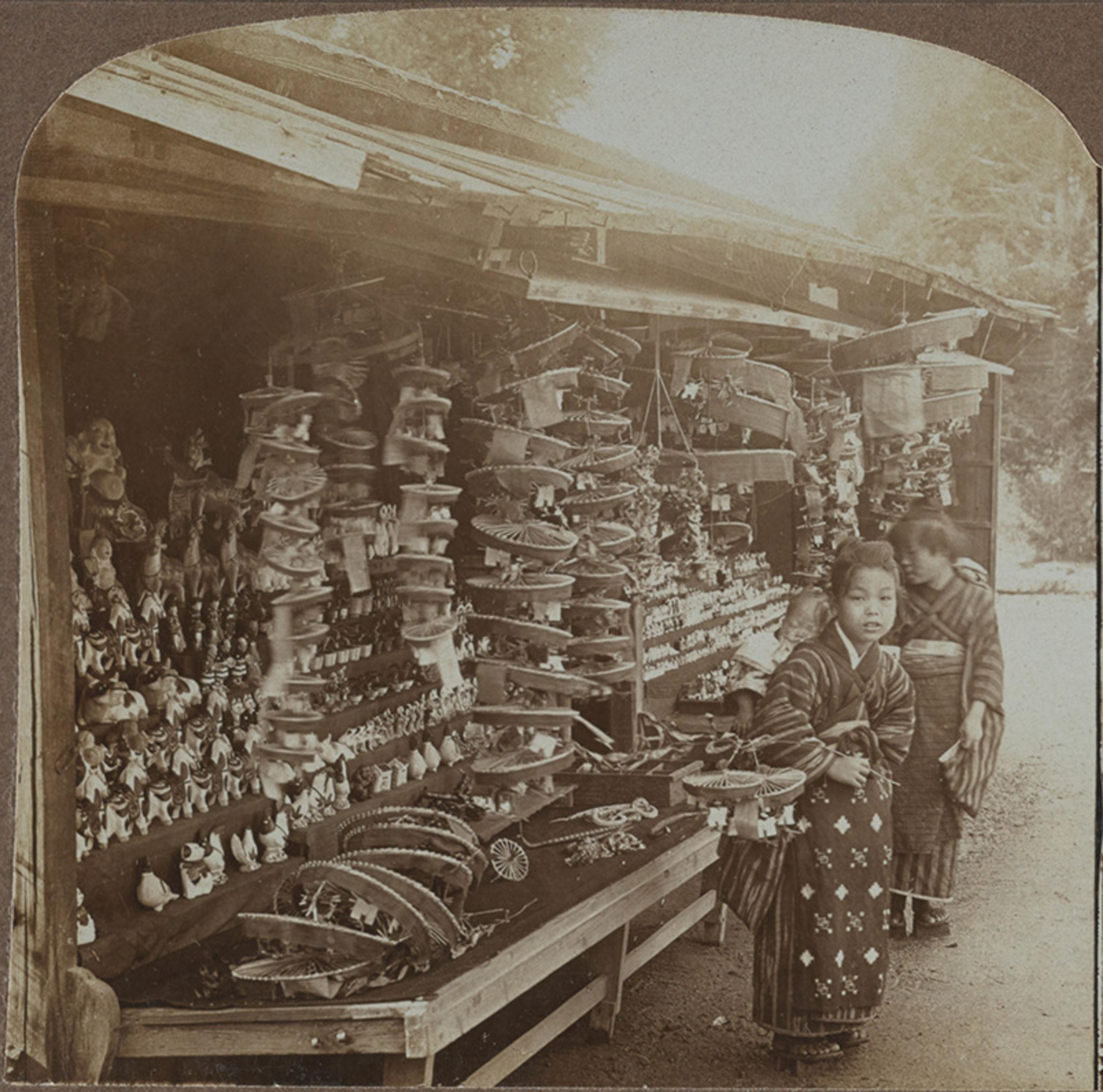 Vintage sepia-toned photo of a market stand in Japan displaying various handicrafts with two children in traditional clothing.