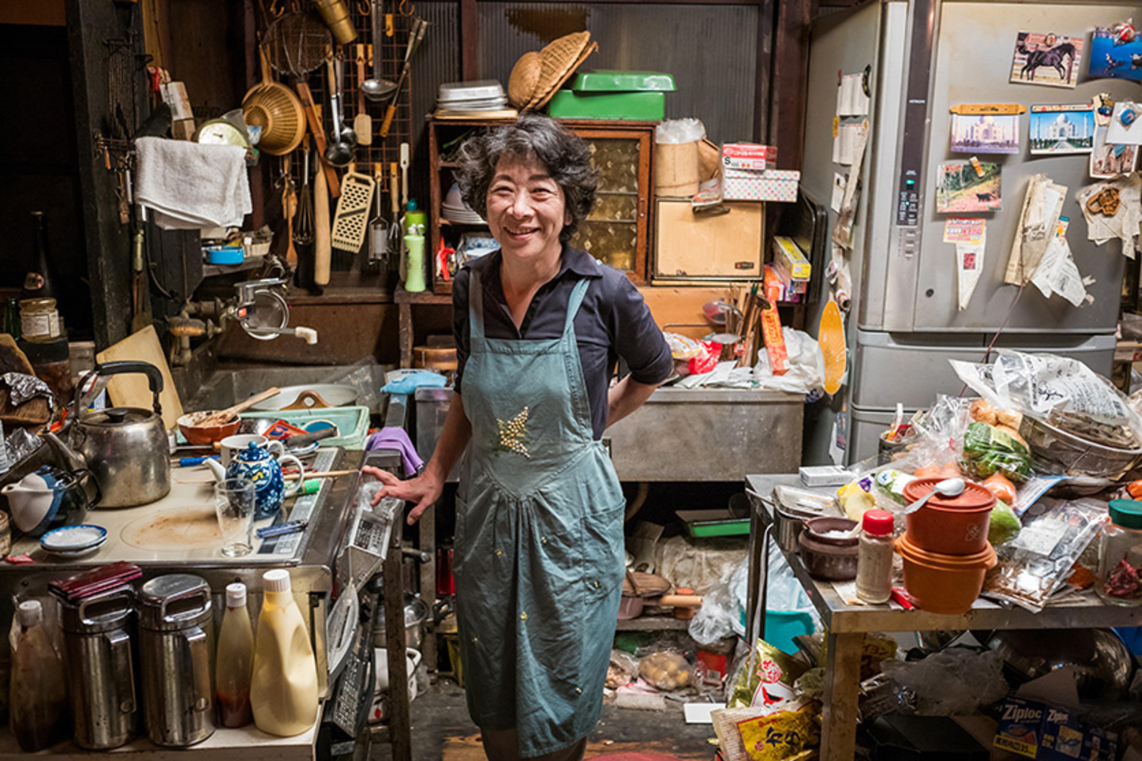 A Japanese woman smiling in a cluttered kitchen with various utensils, groceries and dishes scattered around.