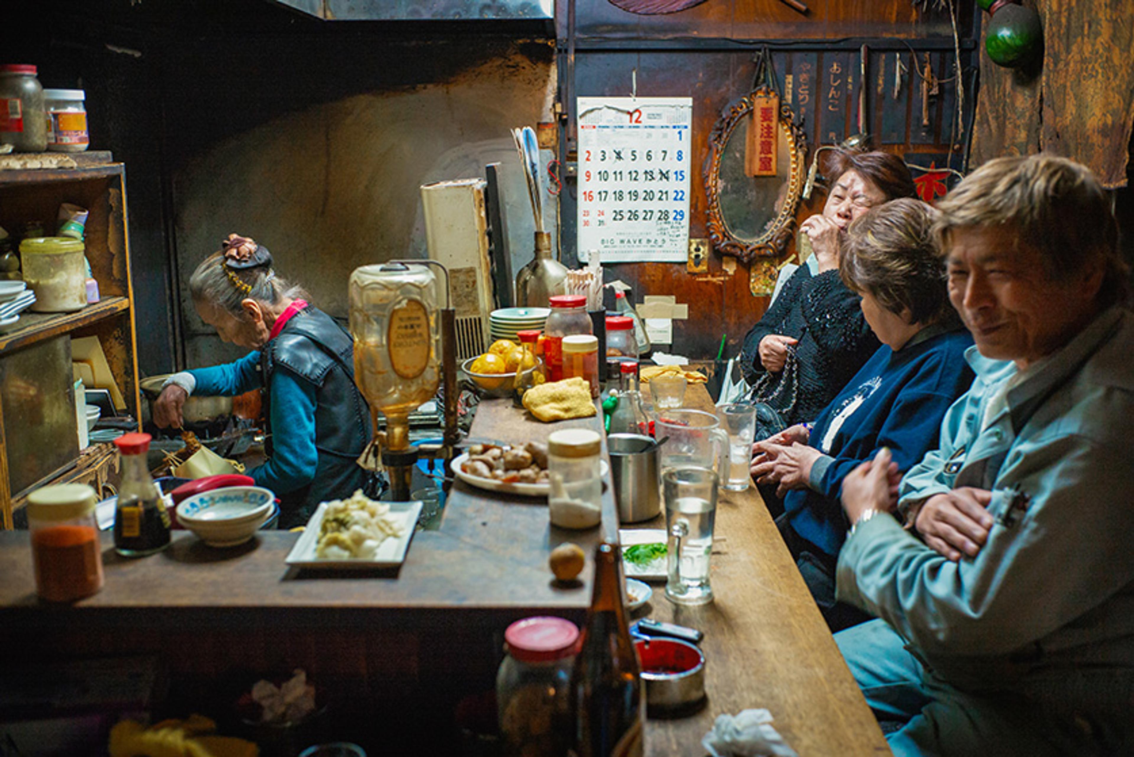 Three people sitting at a cluttered Japanese restaurant counter while an older woman cooks.