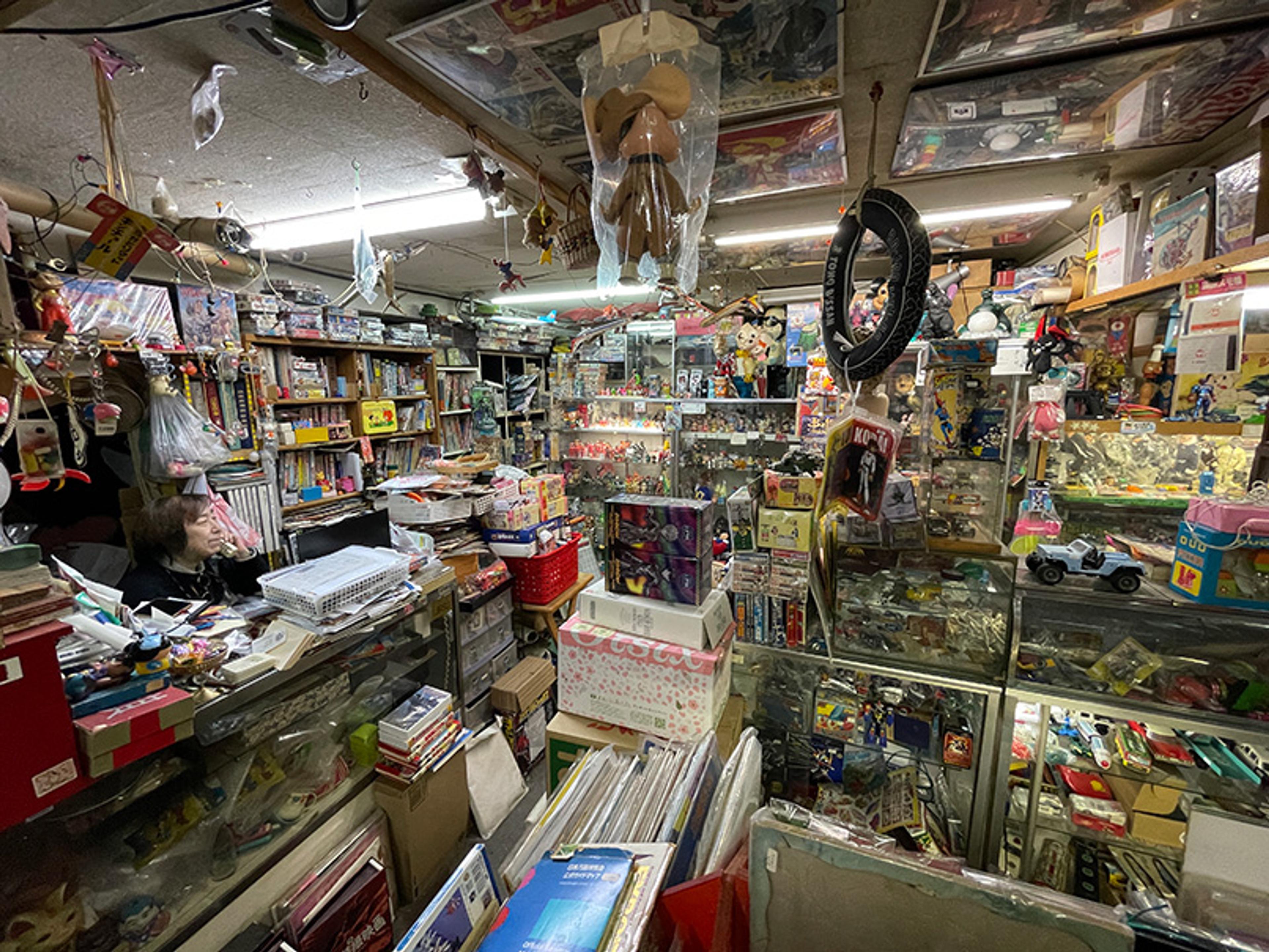 A cluttered, nostalgic toy shop filled with shelves, boxes, action figures, books and a person sitting at a desk.