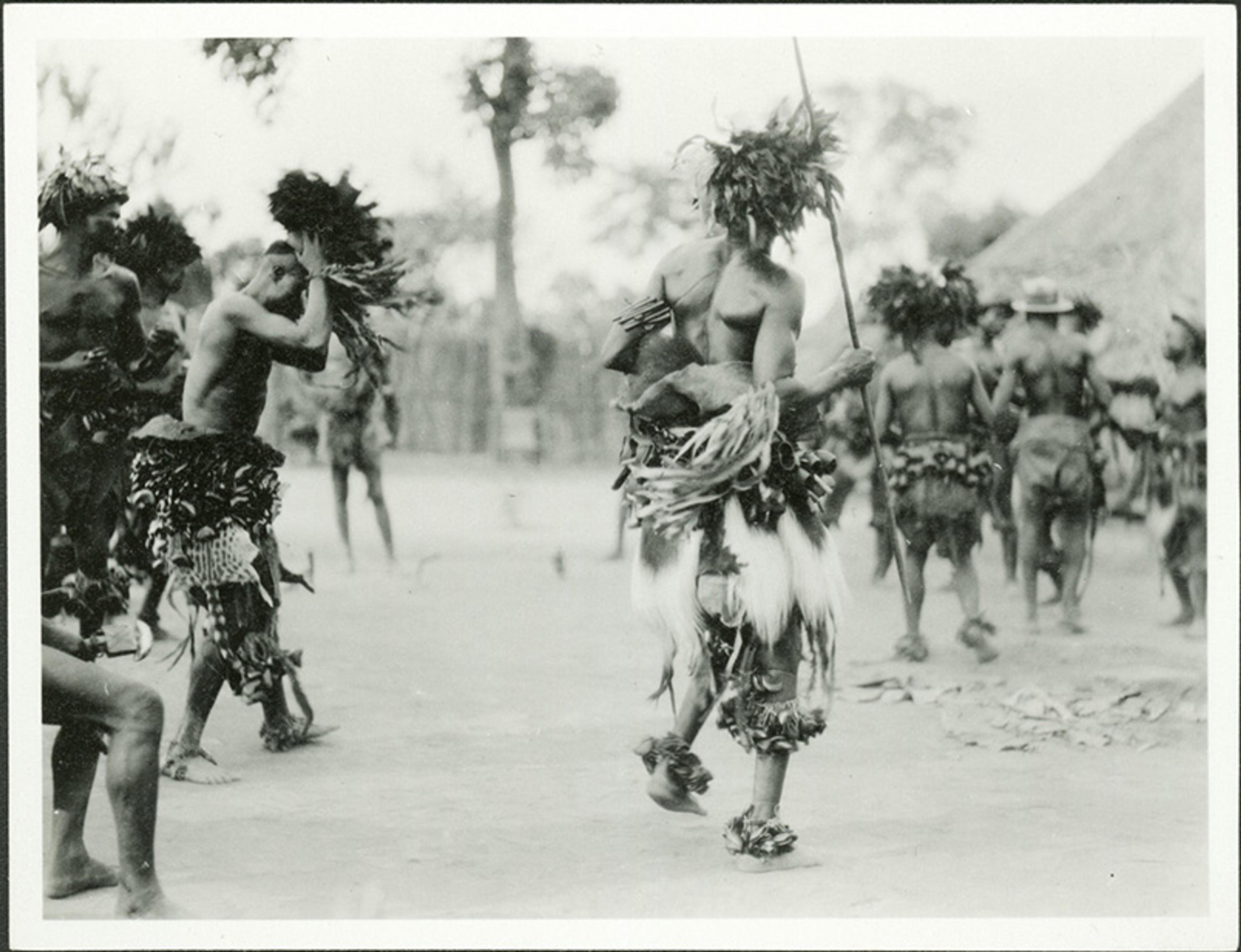 Black and white photo of people in traditional attire dancing outdoors, some holding sticks, with trees in the background.