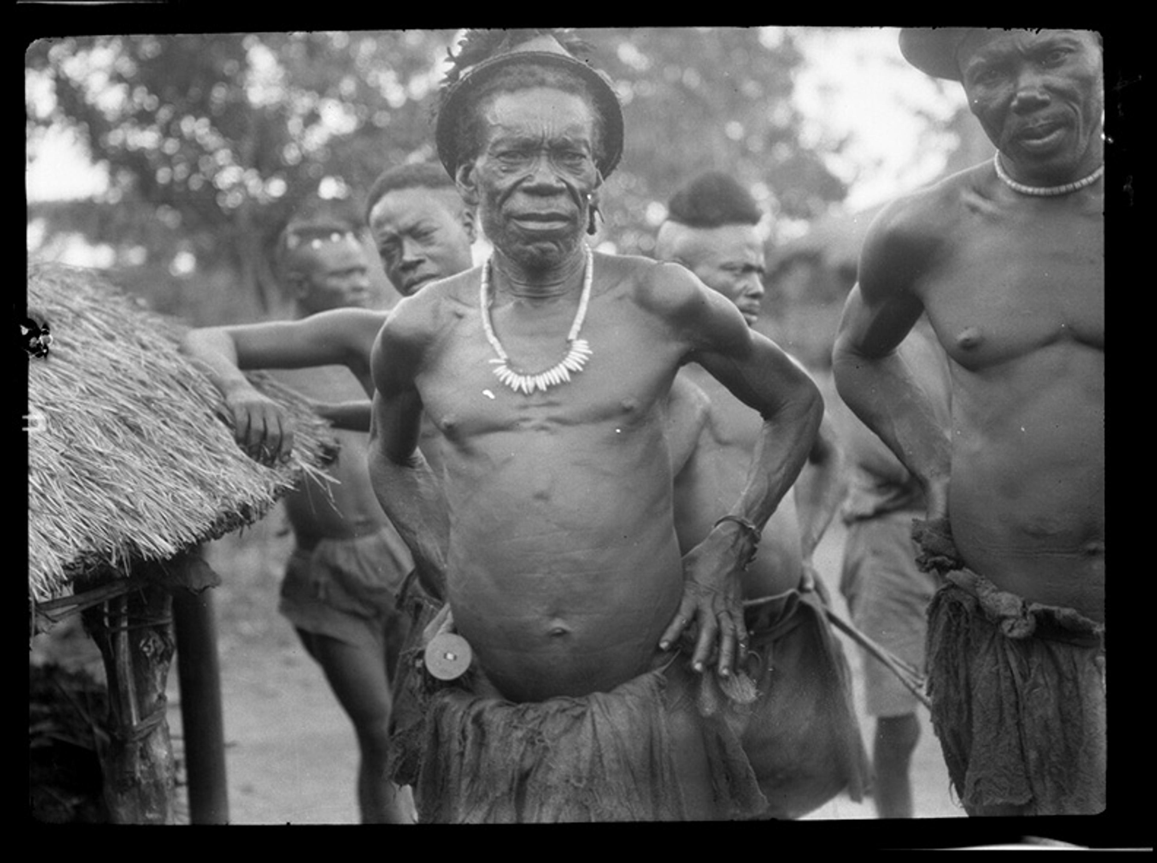 Black and white photo of a group of men in traditional attire outdoors, with huts in the background.