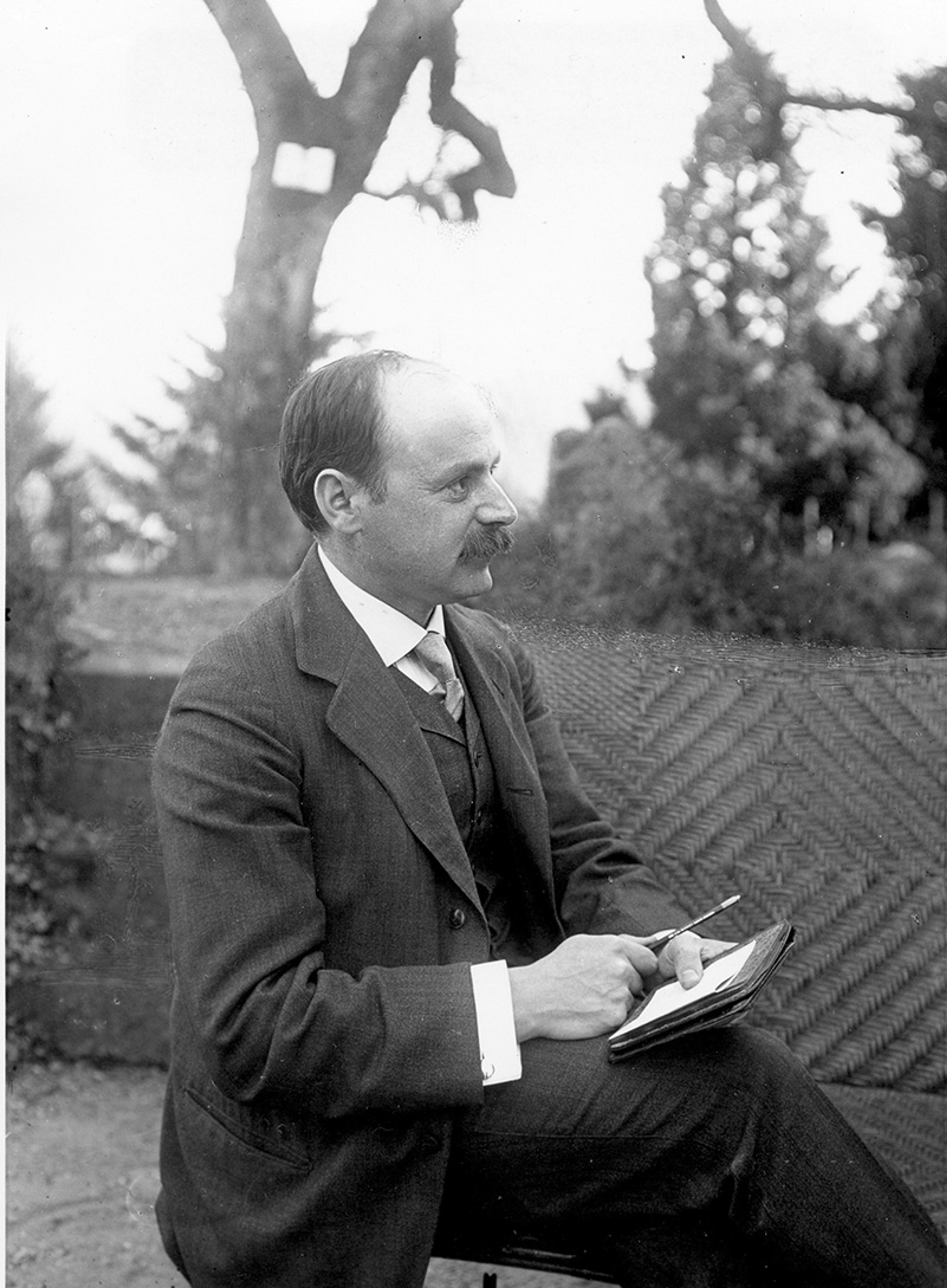 Black and white photo of a man in a suit sitting outdoors holding a pen and paper with trees in the background.