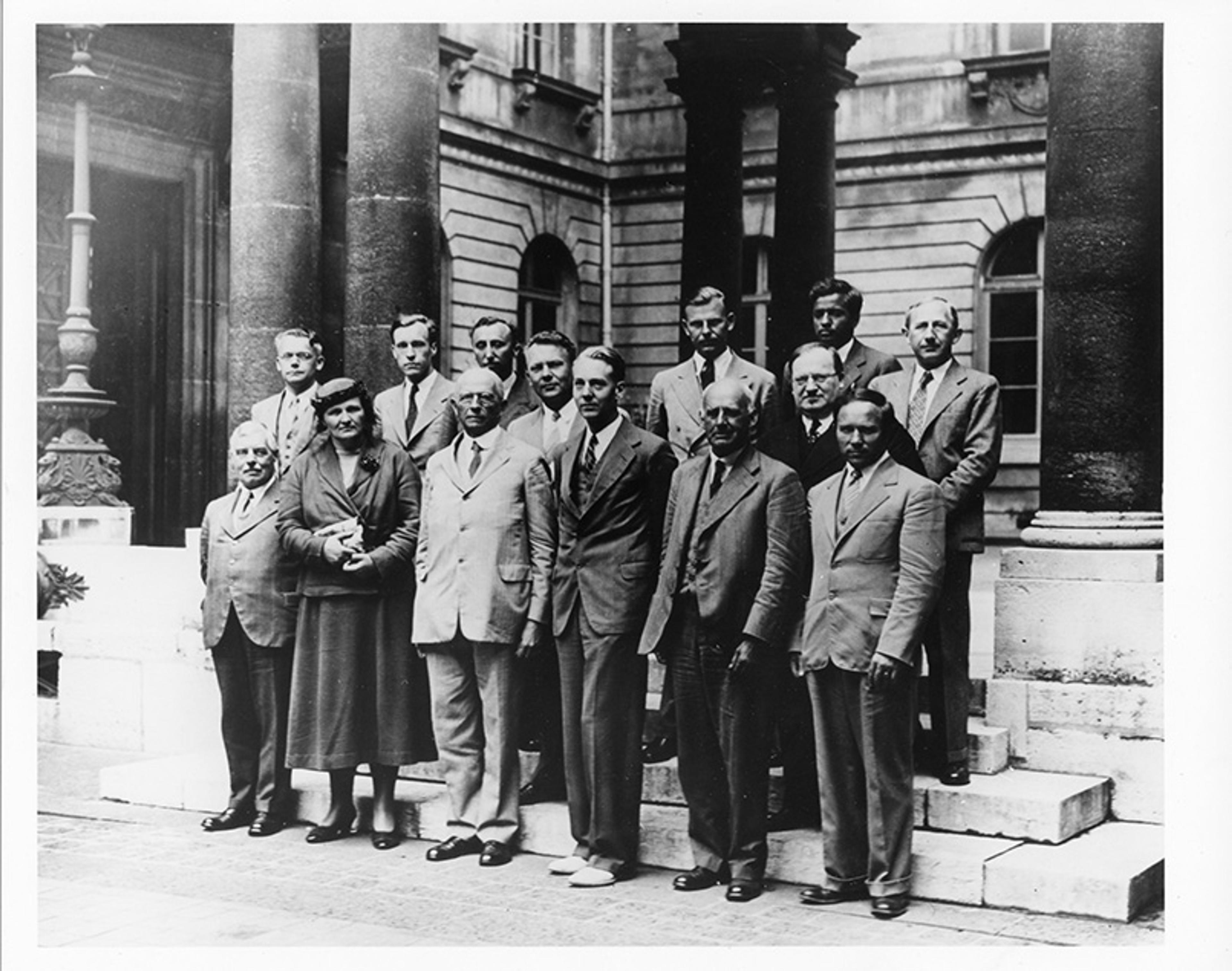 Black and white photo of a group of men and one woman in suits standing on steps with a classical building in the background.