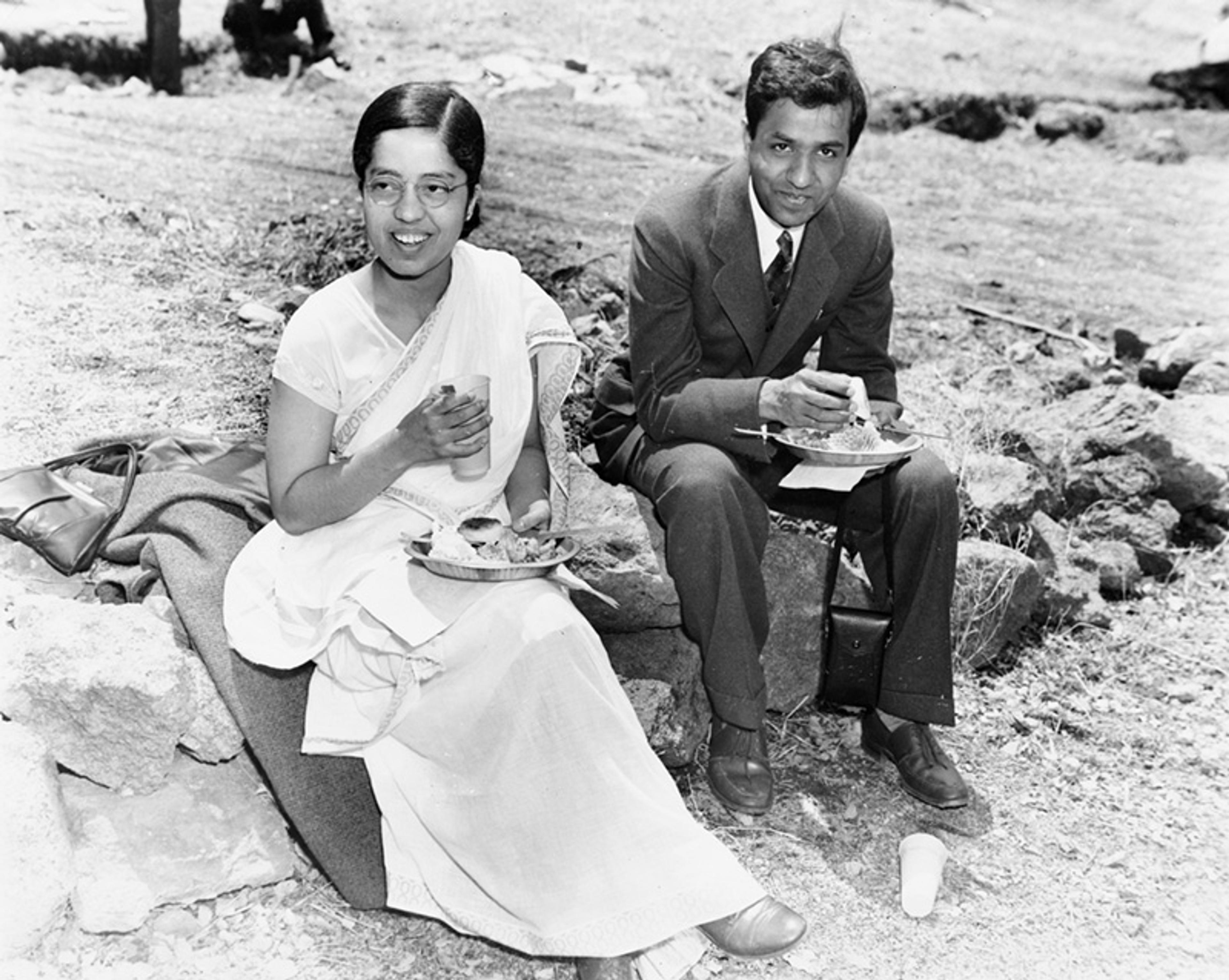 Black and white photo of a woman in a sari and a man in a suit sitting outdoors on rocks eating a meal.