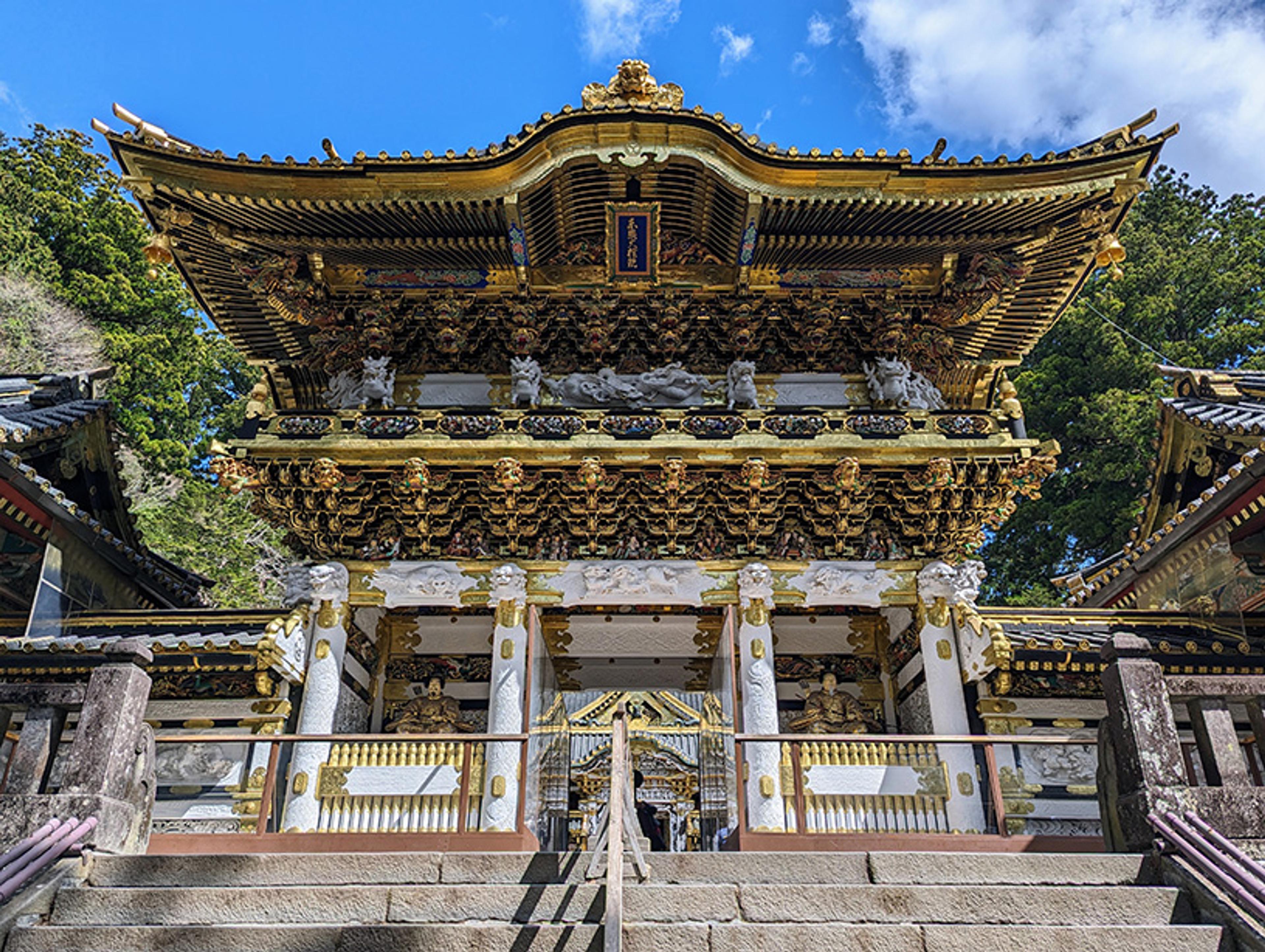 Photo of a richly decorated traditional Japanese temple gate with gold details and ornate carvings, set against a blue sky.