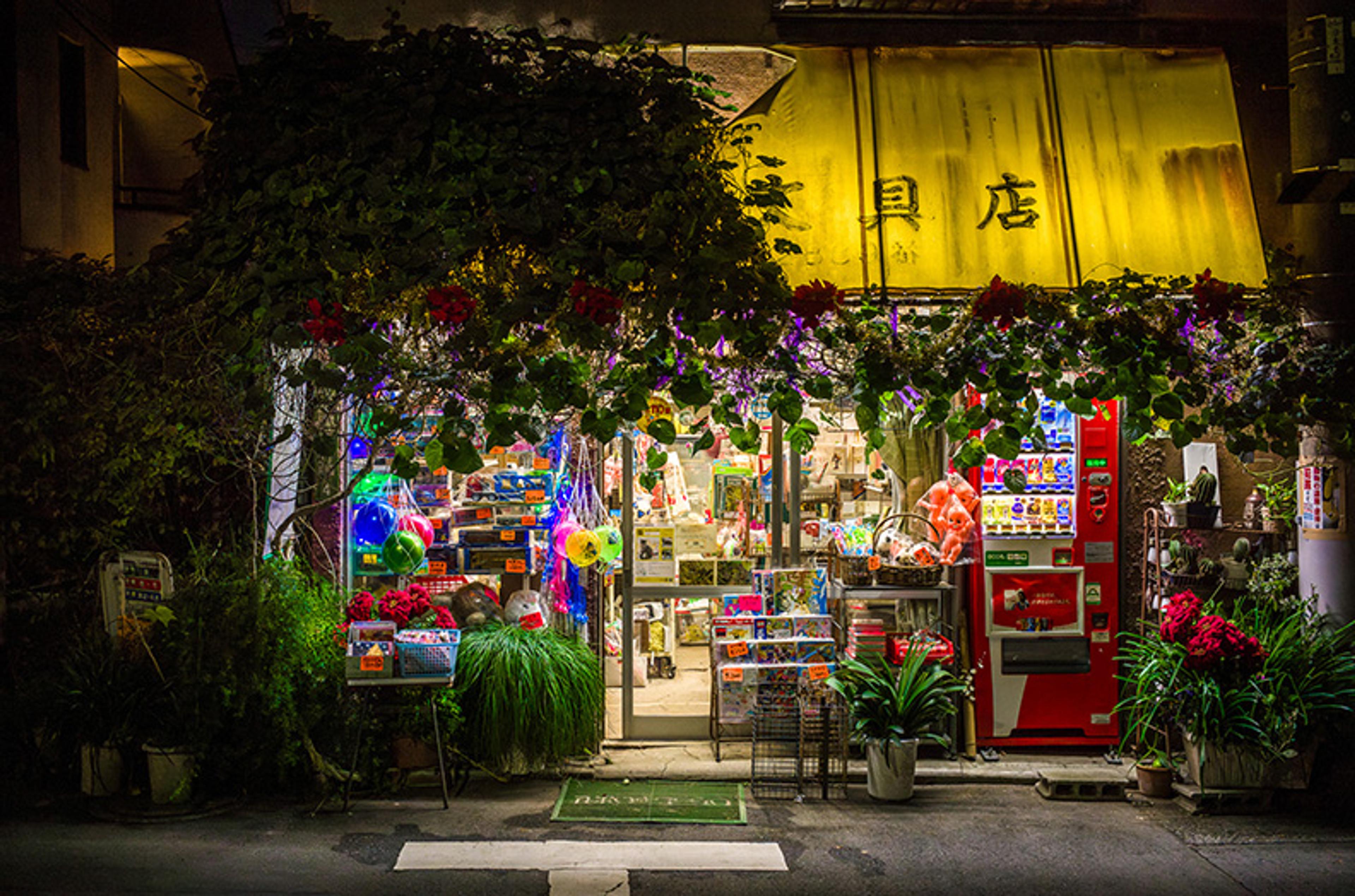 Photo of a small, brightly lit shop at night with a yellow awning, colourful vending machines, plants and flowers outside.