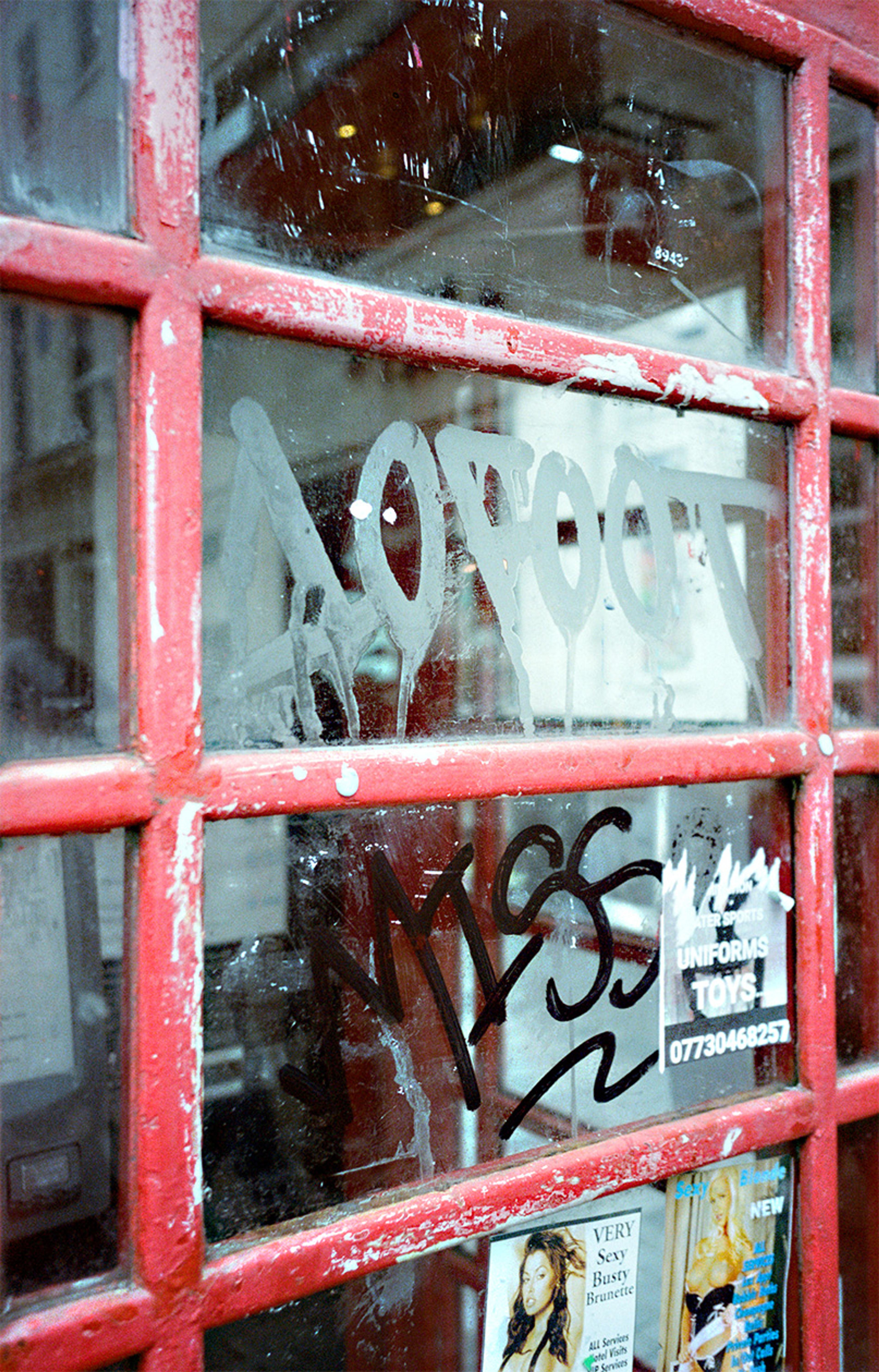 Photo of a red phone box window with graffiti and stickers on the glass, including adverts for adult services.