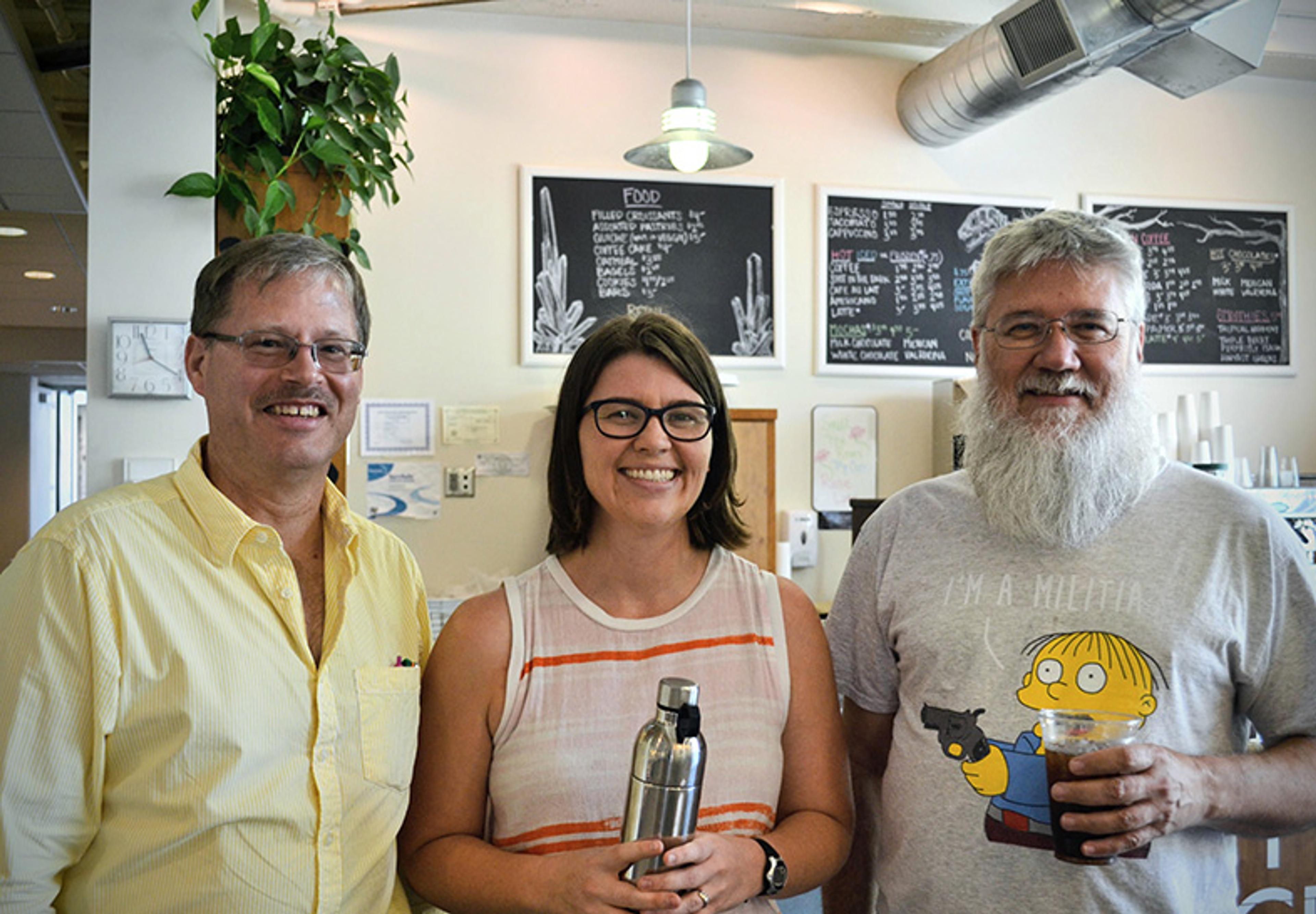 Photo of three smiling people in a café with menus in the background, one holding a drink and one with a water bottle.