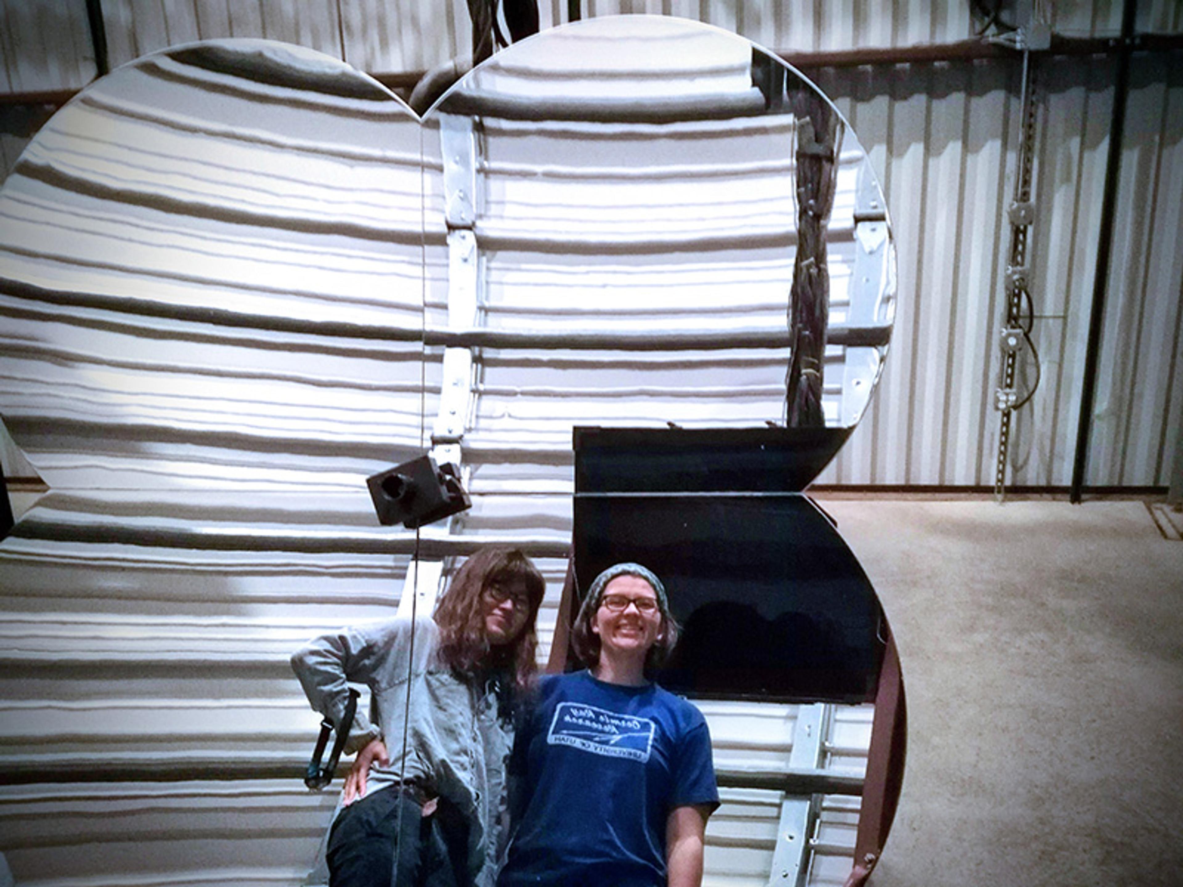 Photo of two people smiling in front of a large mirrored object inside a warehouse-like setting.