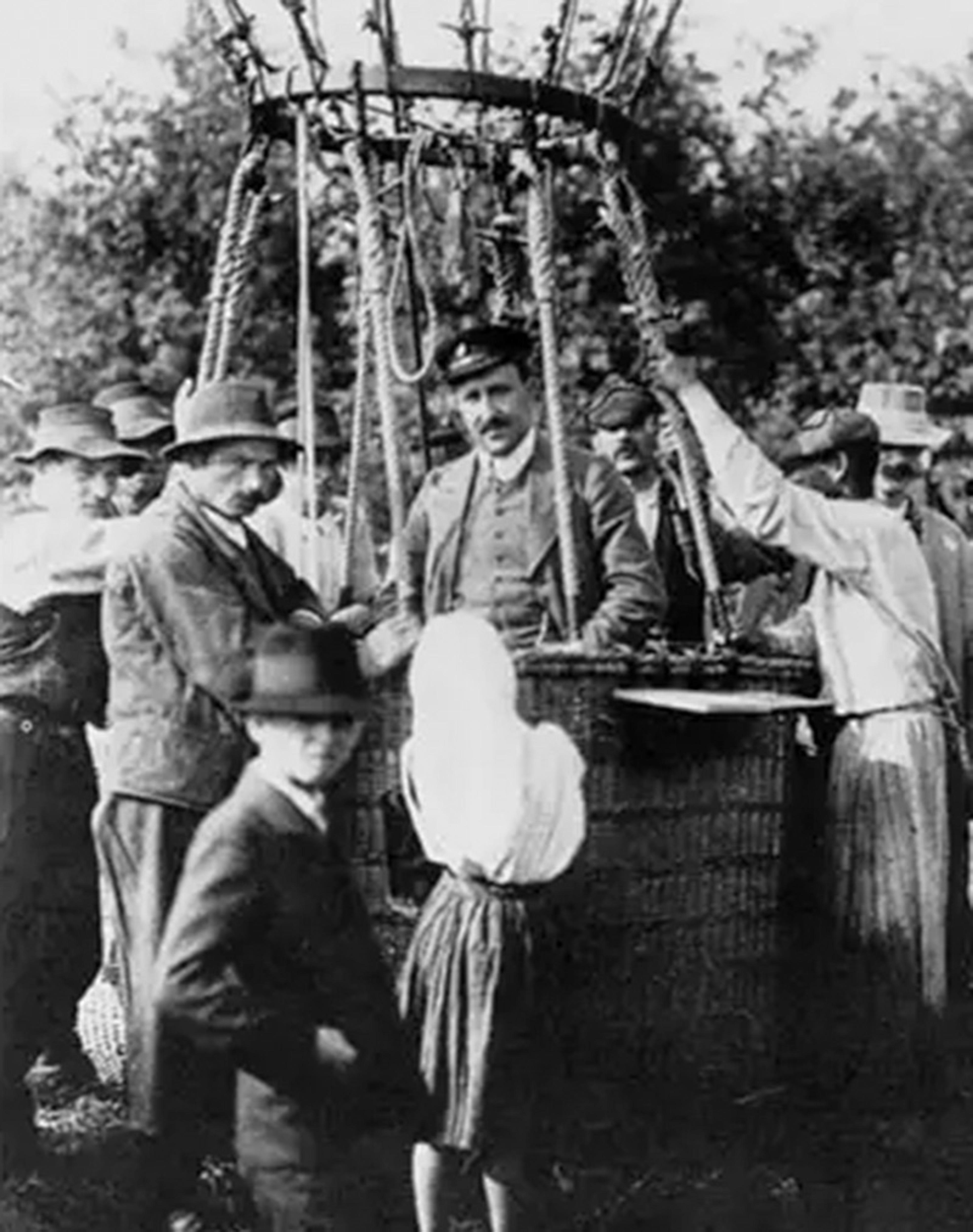 Vintage photo of a crowd gathered around a hot air balloon basket in a park, with people dressed in early 20th-century attire.