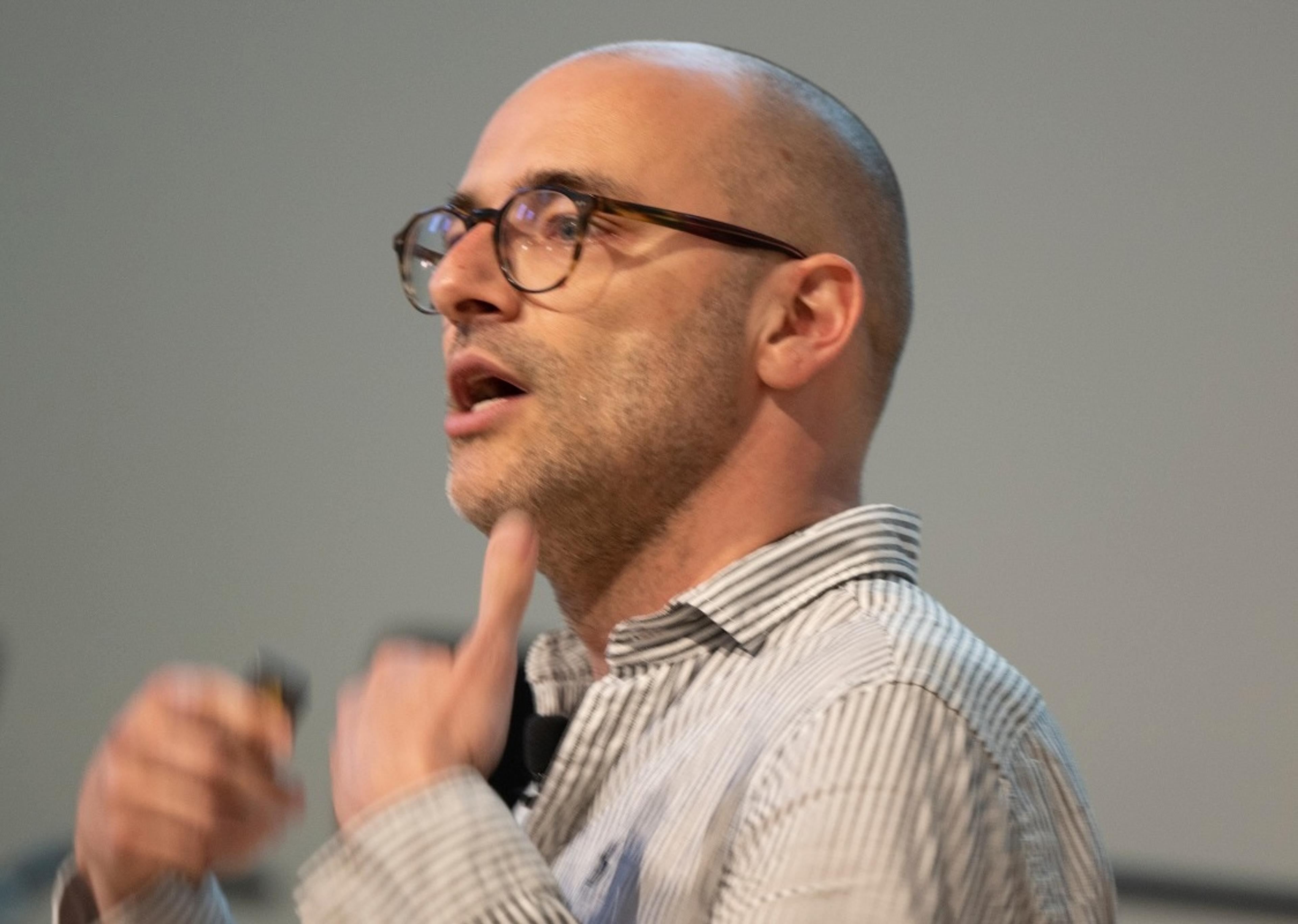 Photo of a bald man wearing glasses and a striped shirt speaking with a hand gesture against a plain background.