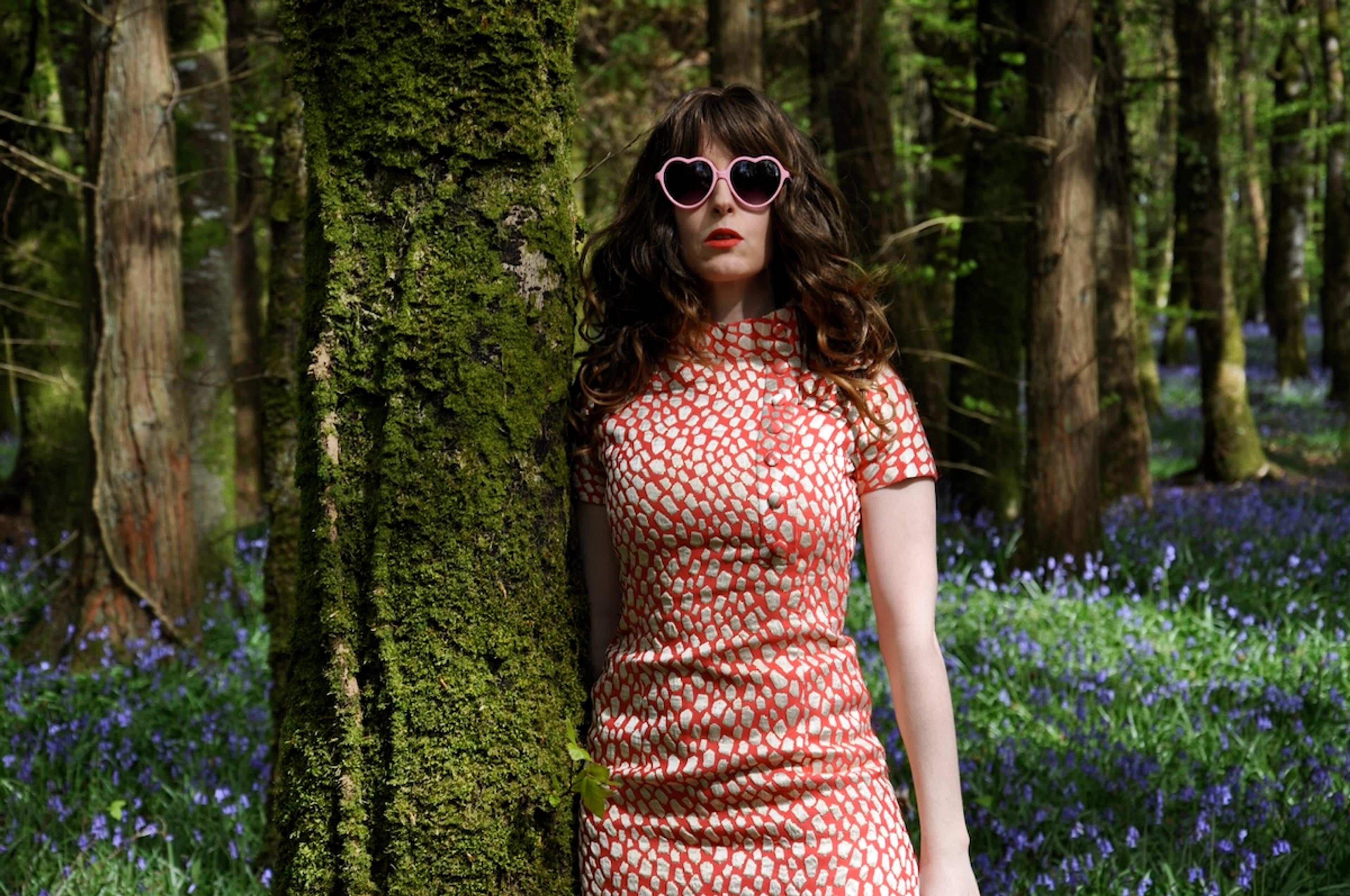 Photo of a woman in a red dress and heart-shaped sunglasses in a forest with purple flowers and moss-covered trees.