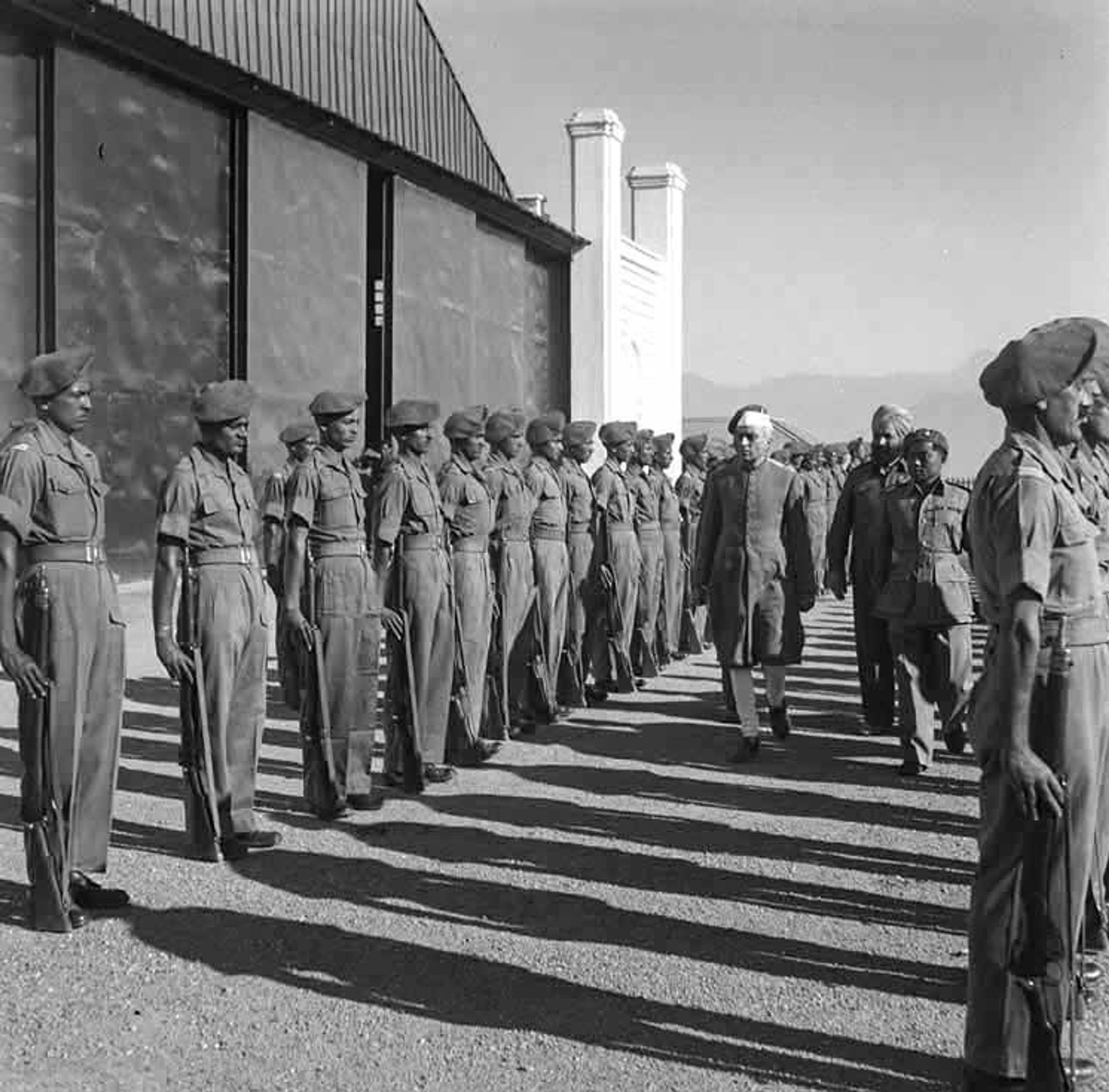 Black-and-white photo of soldiers in uniform standing in formation as officials walk past outside a large building.