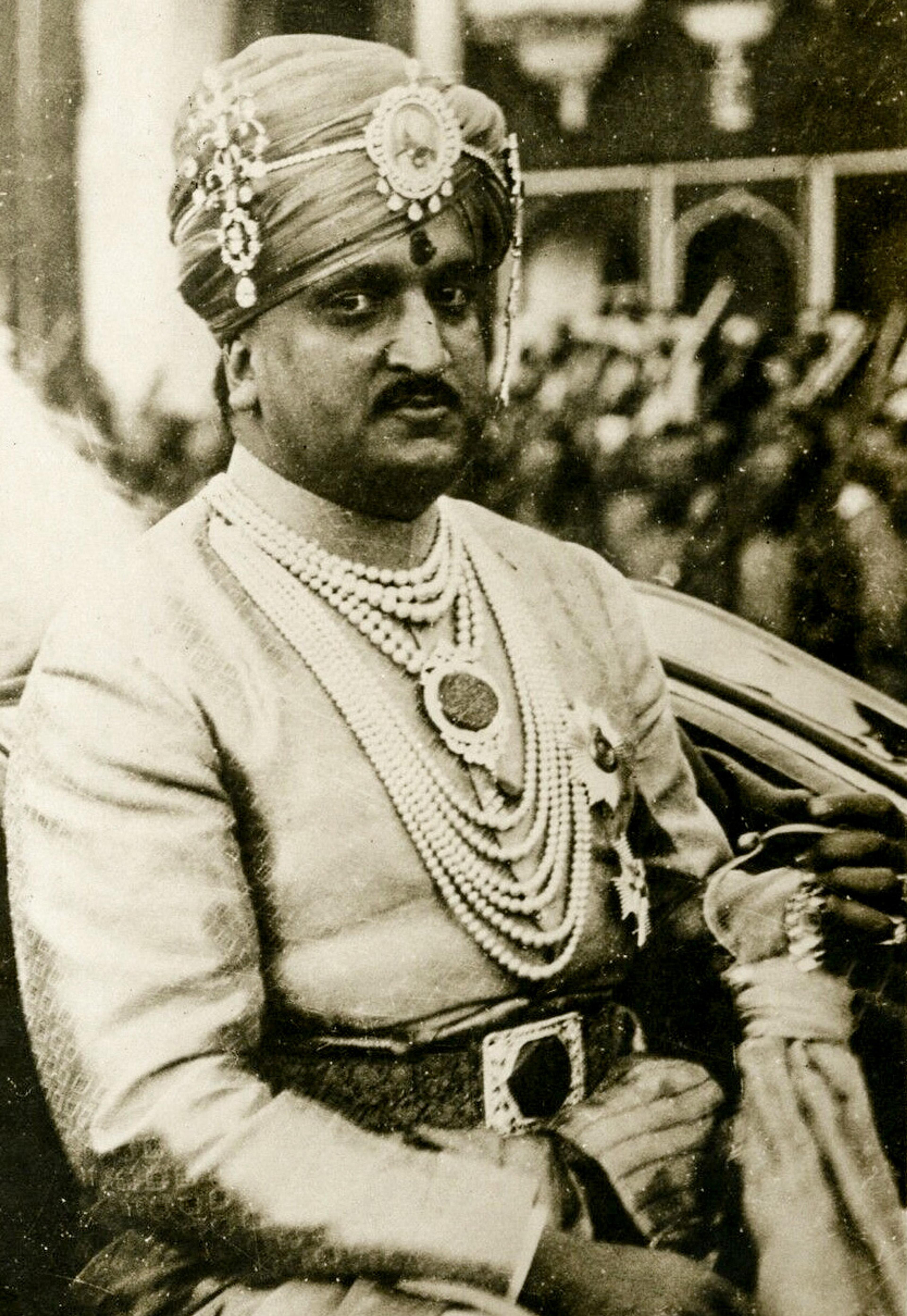 Black and white photo of a man in royal attire with a turban and jewellery sitting in a car.