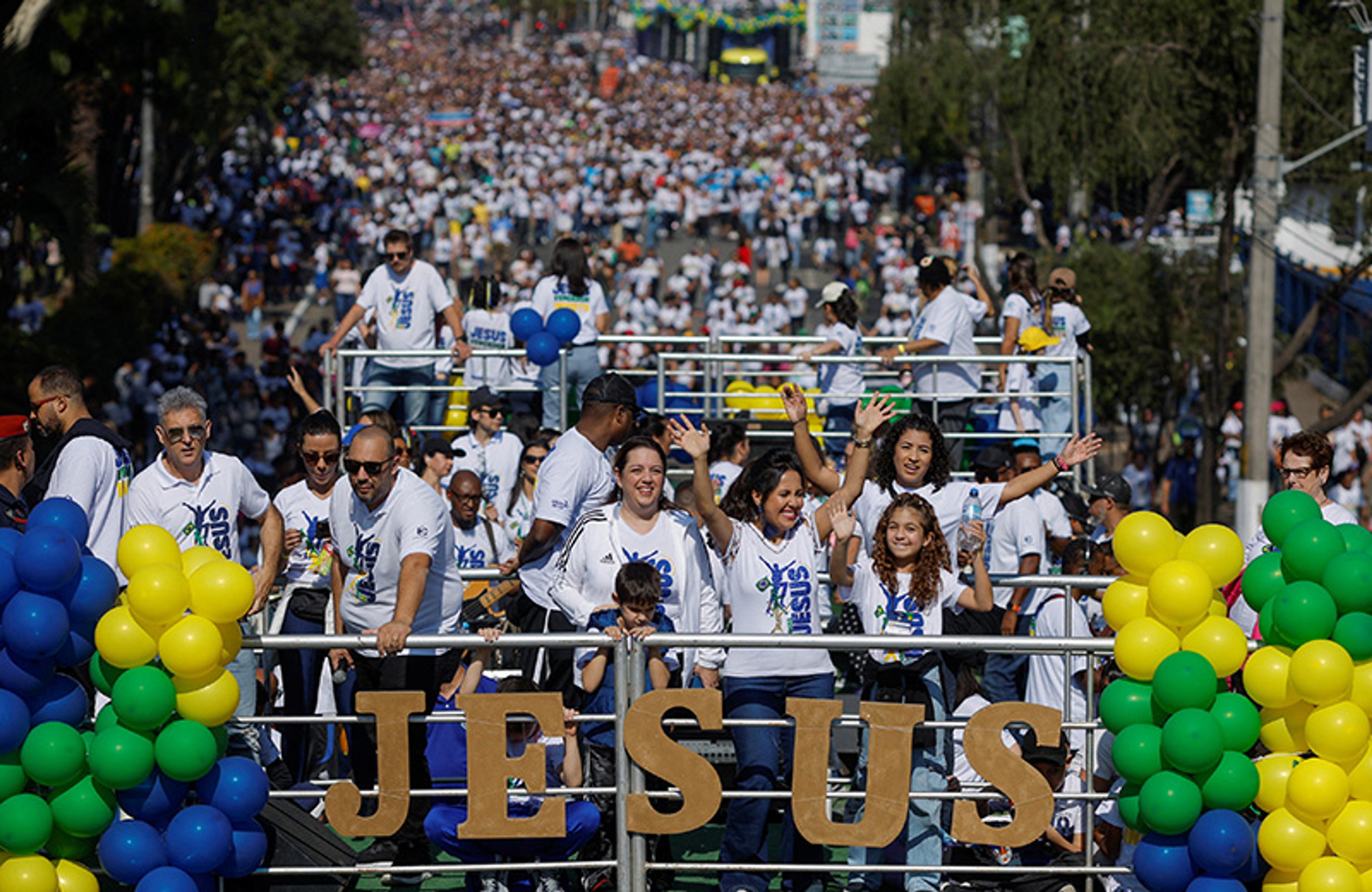 A large crowd at a parade with people in white shirts on a float with JESUS sign and colourful balloons.