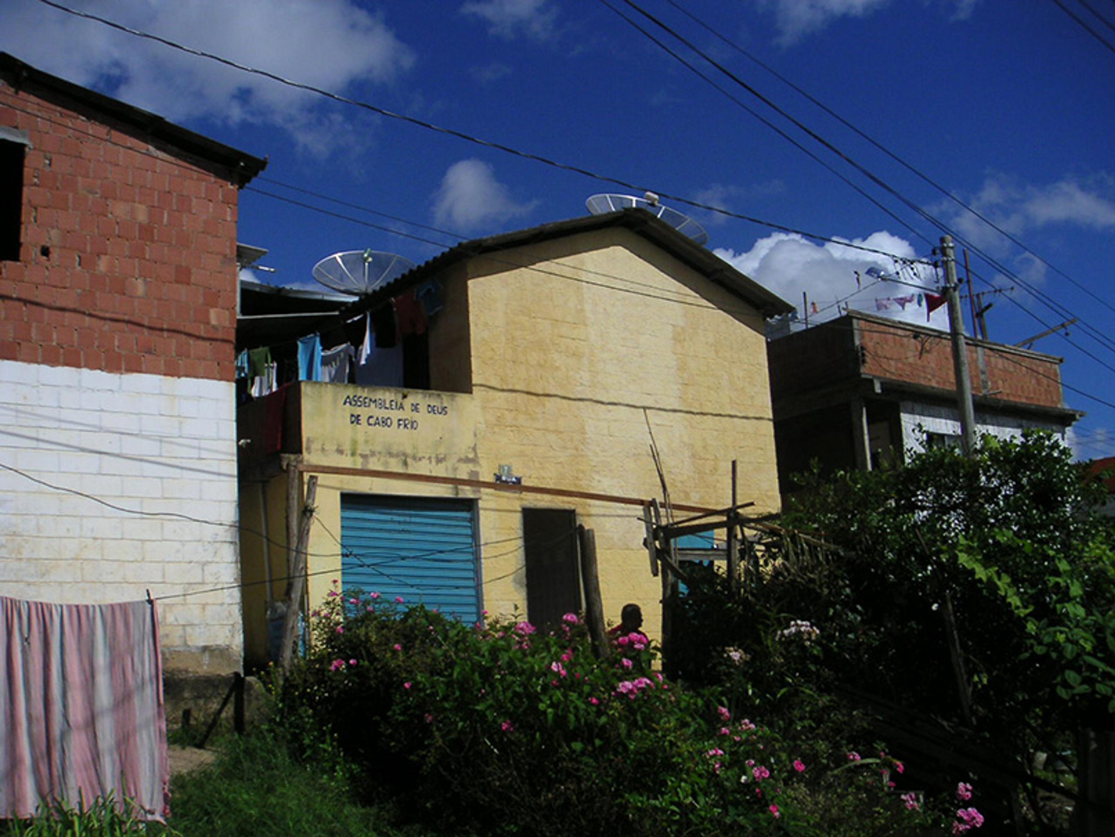 A rustic building with clotheslines and satellite dishes, with buildings close by on either side and bushes in the foreground under a blue sky.