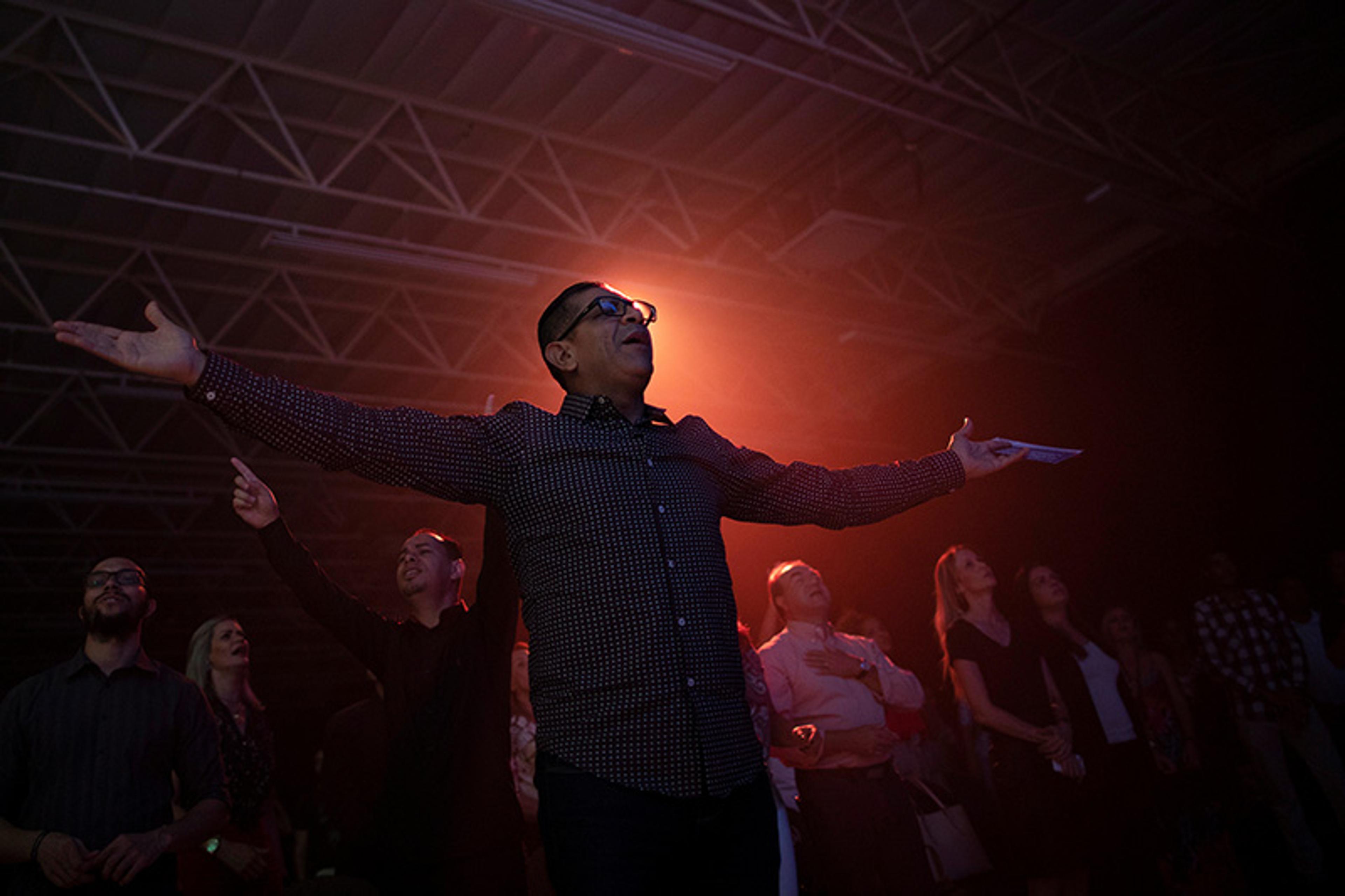 A man with arms outstretched in a dimly lit room filled with people, illuminated by orange light.