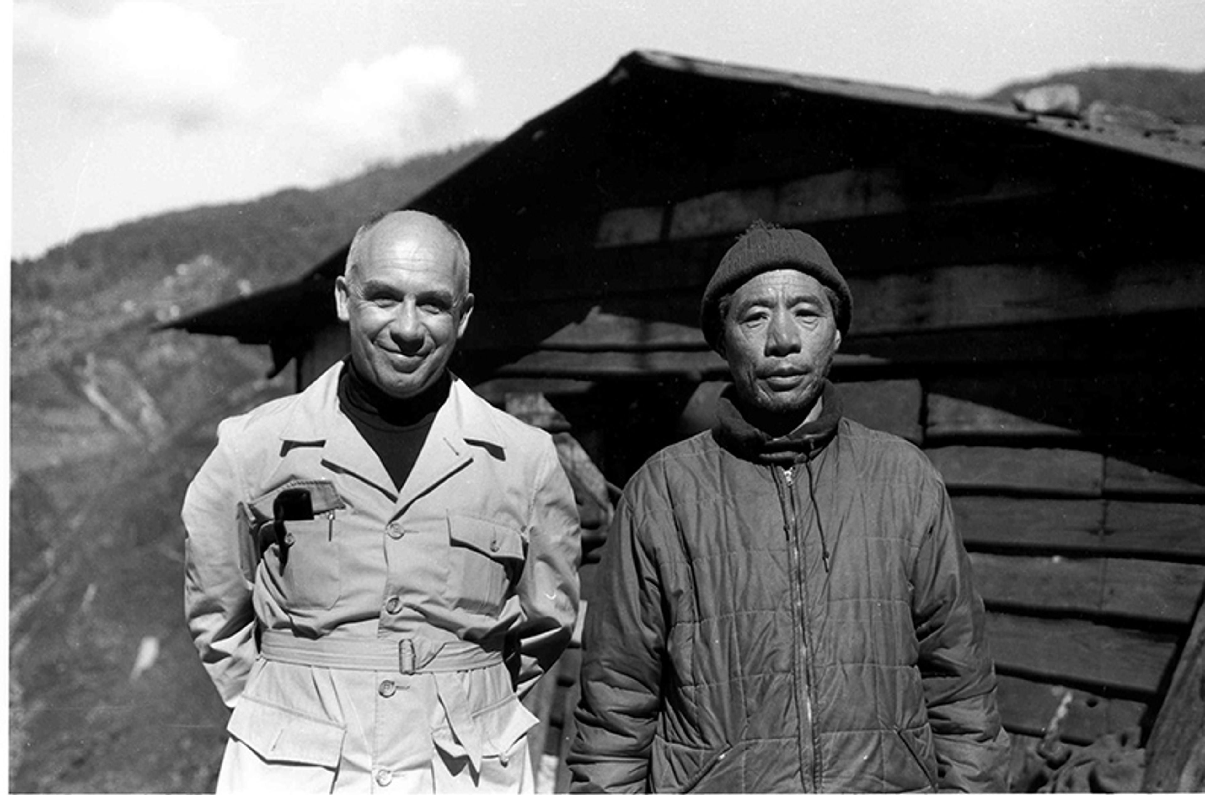 Black and white photo of an American man and a Tibetan man in front of a wooden cabin in a mountainous area.