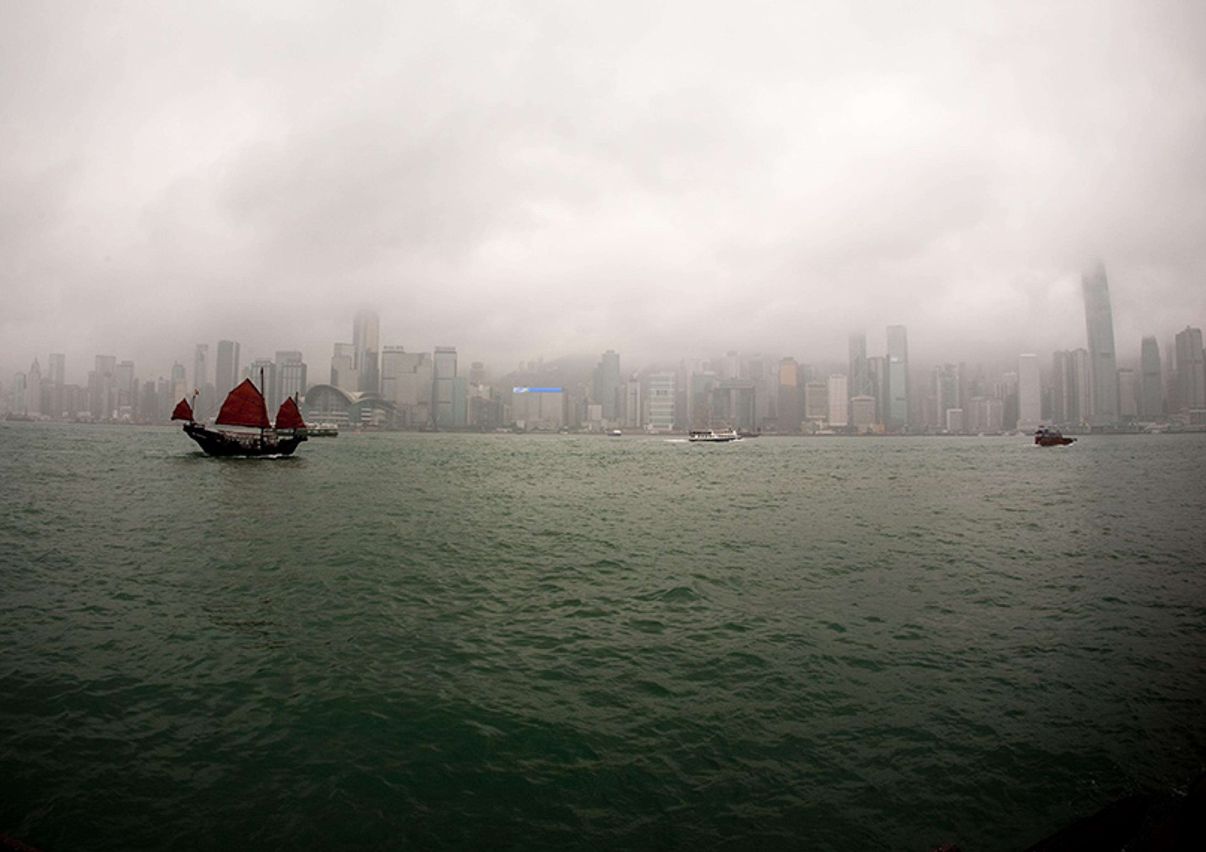 A foggy city skyline with red-sailed junks on the water in the foreground, under an overcast sky.