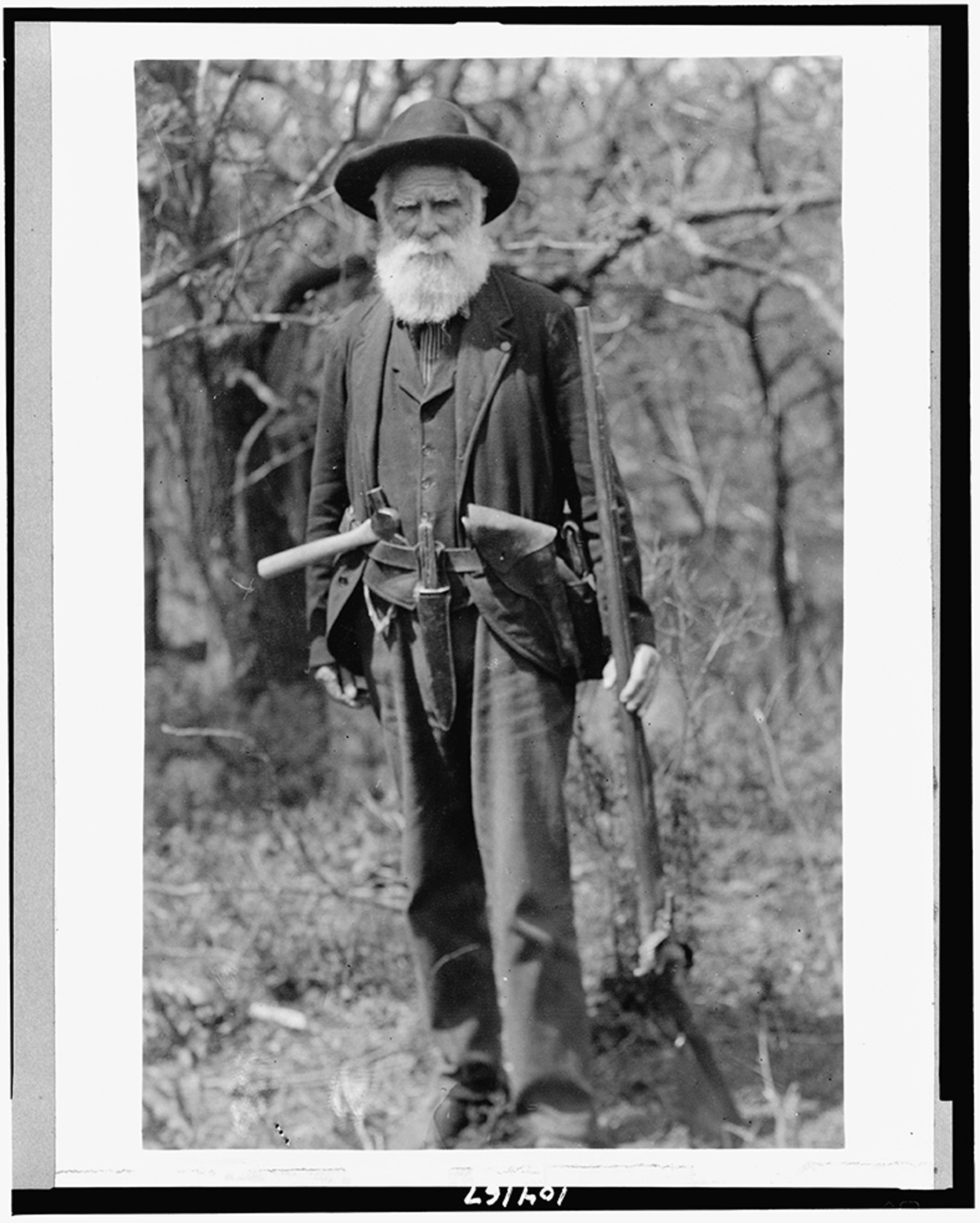 Black and white photo of an elderly bearded man in a hat holding a rifle standing in a wooded area.