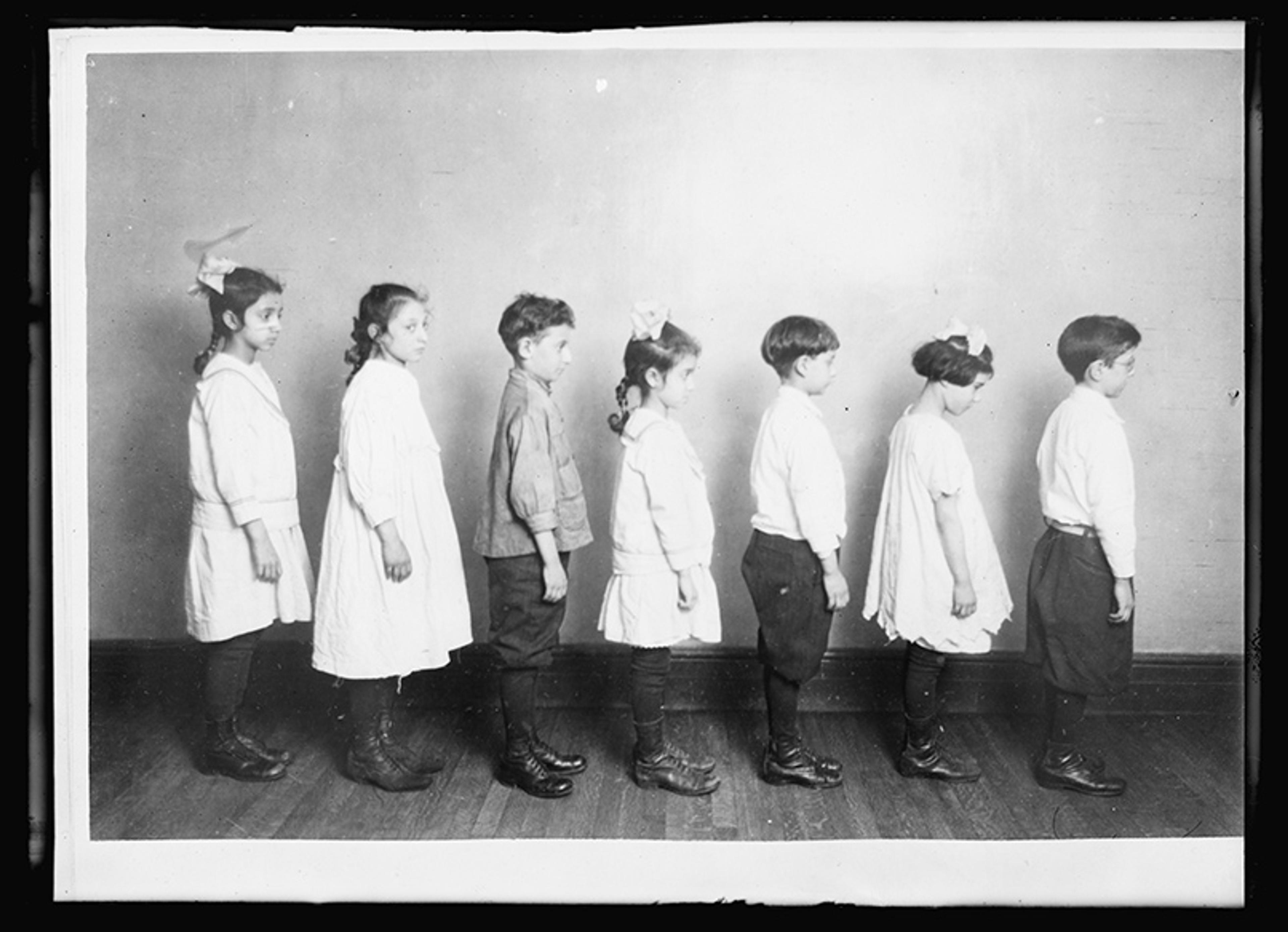 Black and white photo of seven children standing in a line side by side against a plain wall, dressed in early 20th century attire.