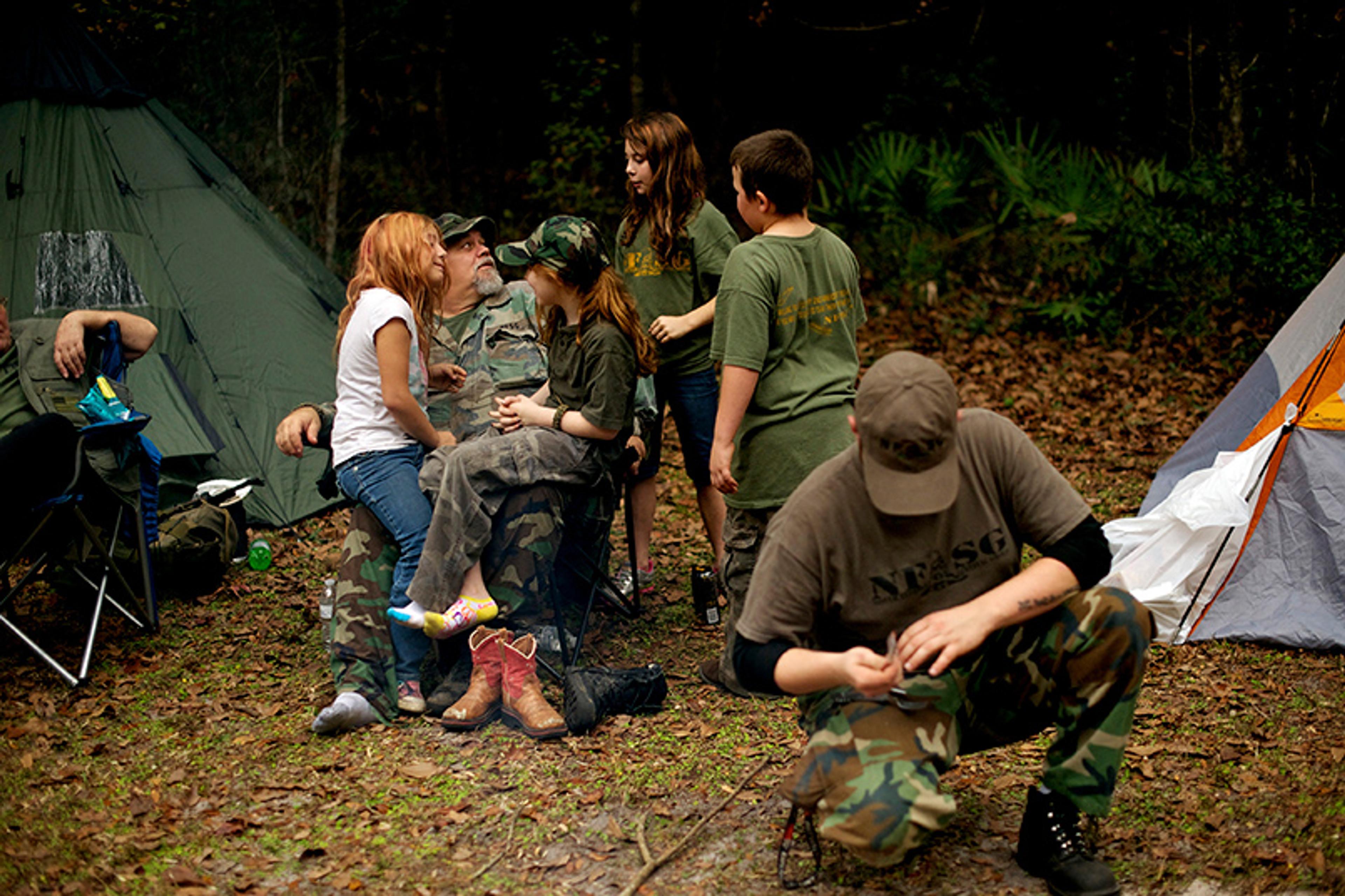 People in camouflage clothing at a campsite with tents in a wooded area. They appear to be preparing or relaxing.