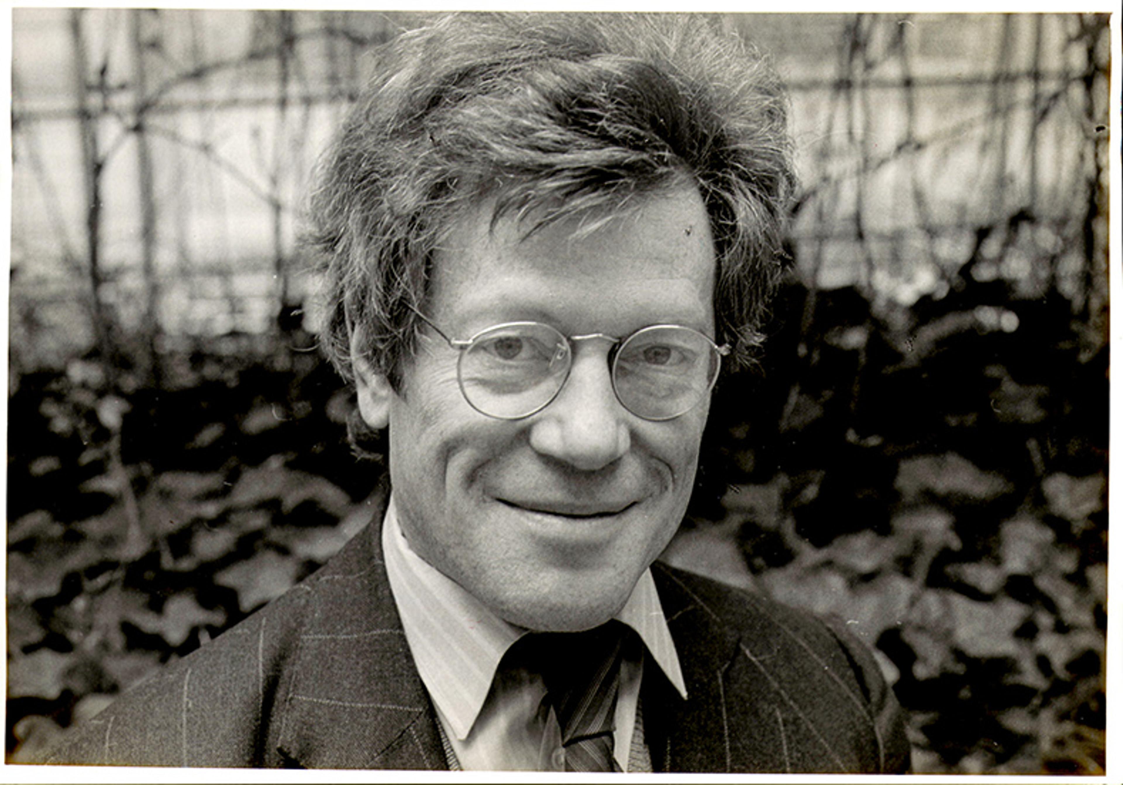 Black and white photo of a man in glasses and a suit smiling outdoors with a blurred leafy background.