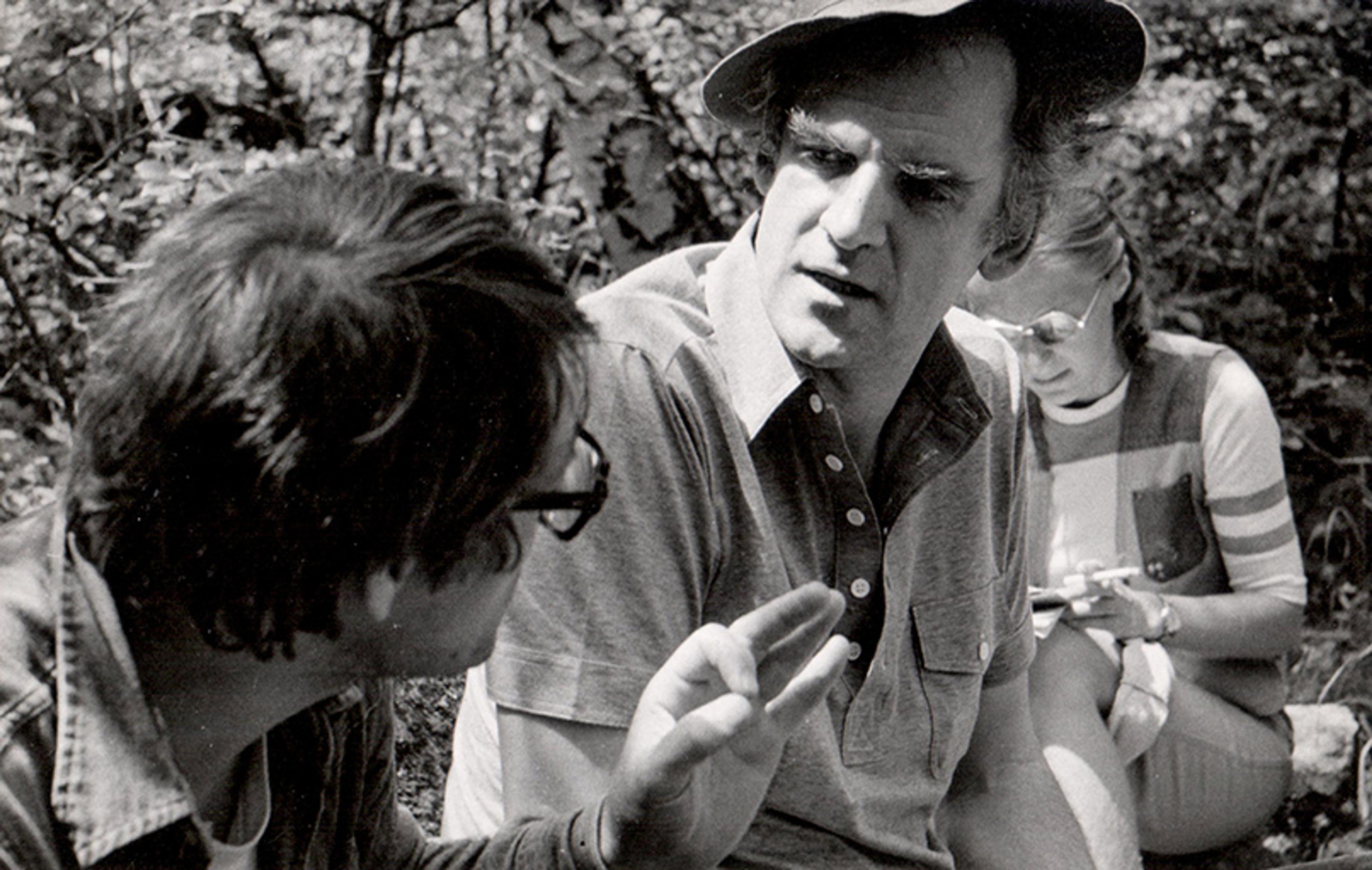 Black and white photo of three people sitting outdoors; two men in conversation, a woman behind with documents in her hands.