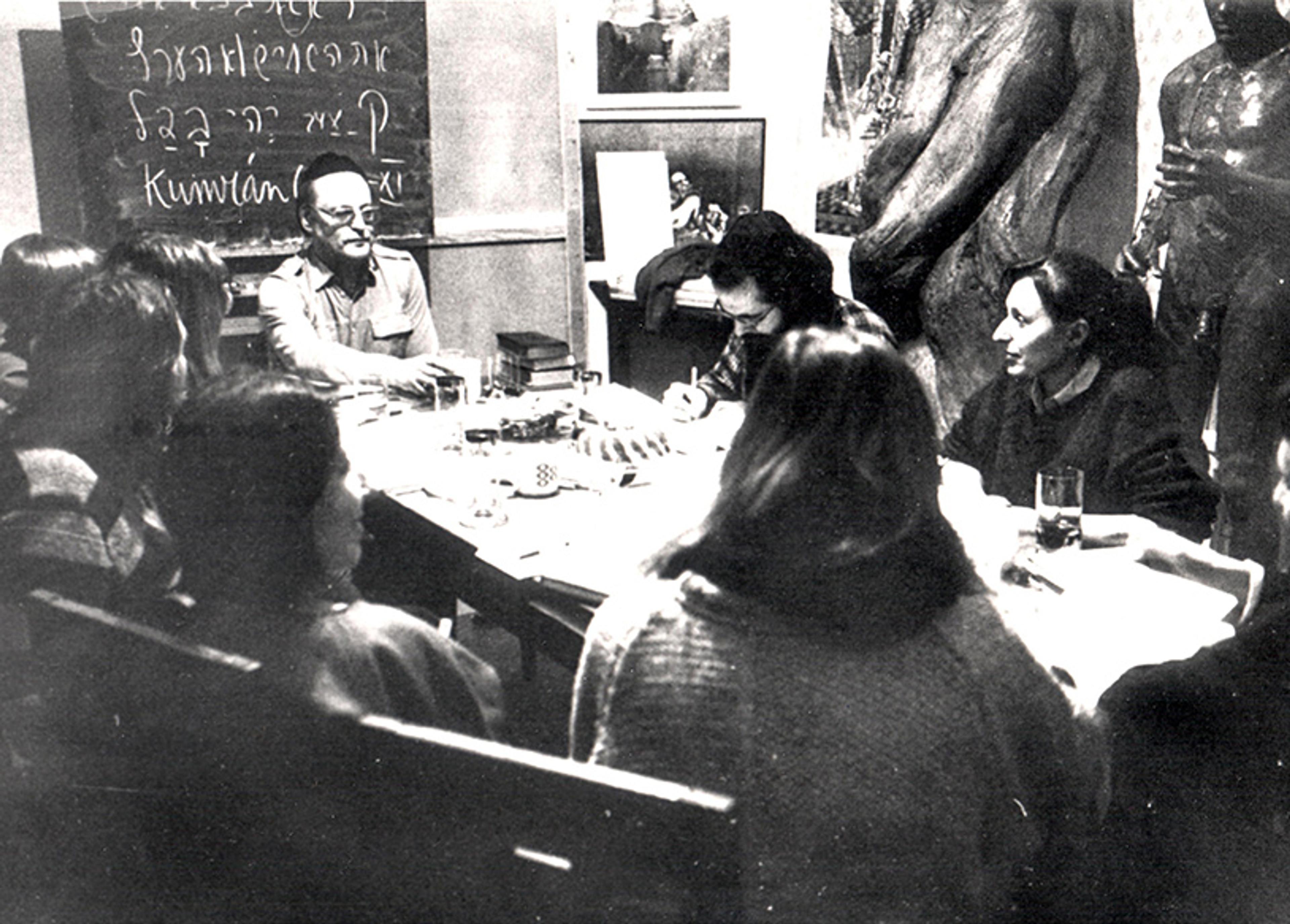 Black and white photo of a group of people gathered around a table with notebooks and drinks in a classroom setting.