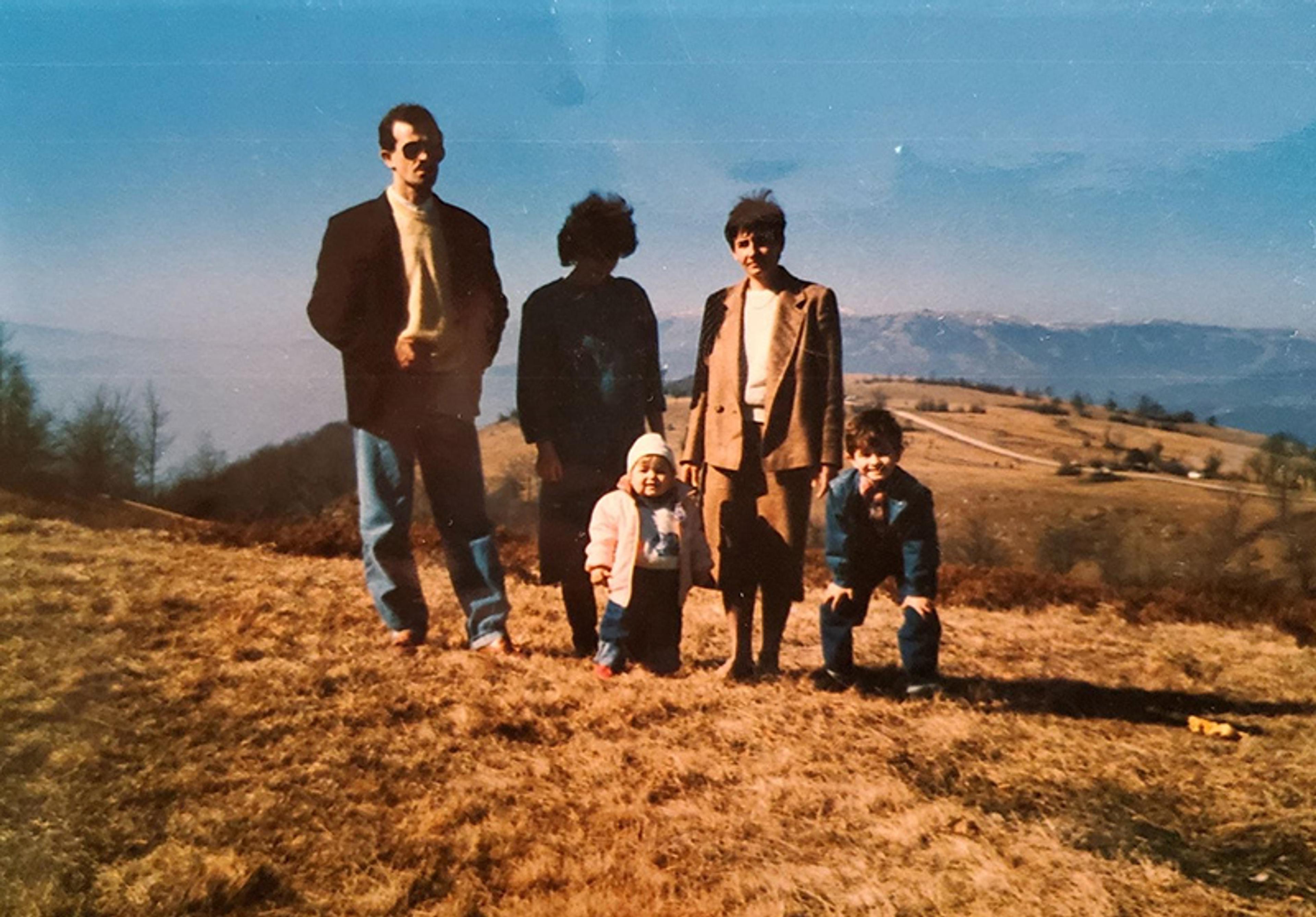 Photo of three adults and two small children on a grassy hill with a mountain backdrop under a clear blue sky.