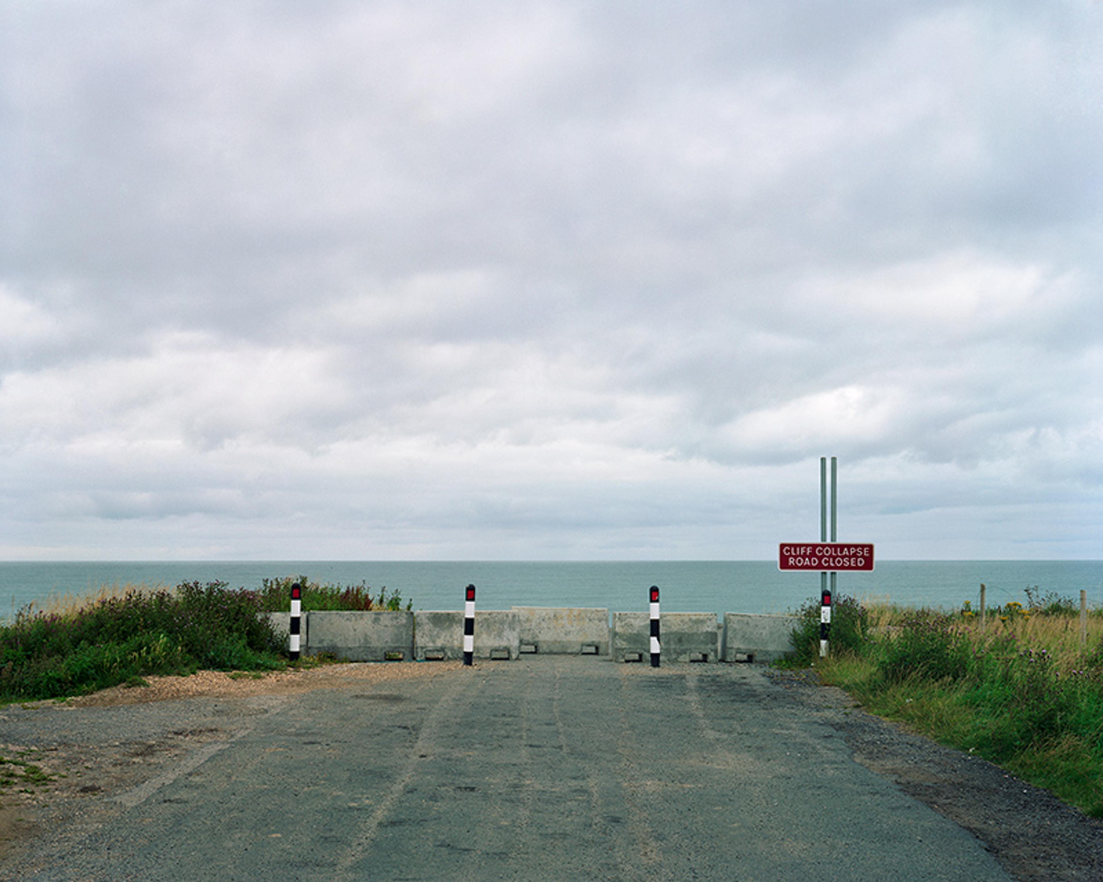 A blocked road leading to the sea, with a “Cliff Collapse Road Closed” sign, under a cloudy sky.